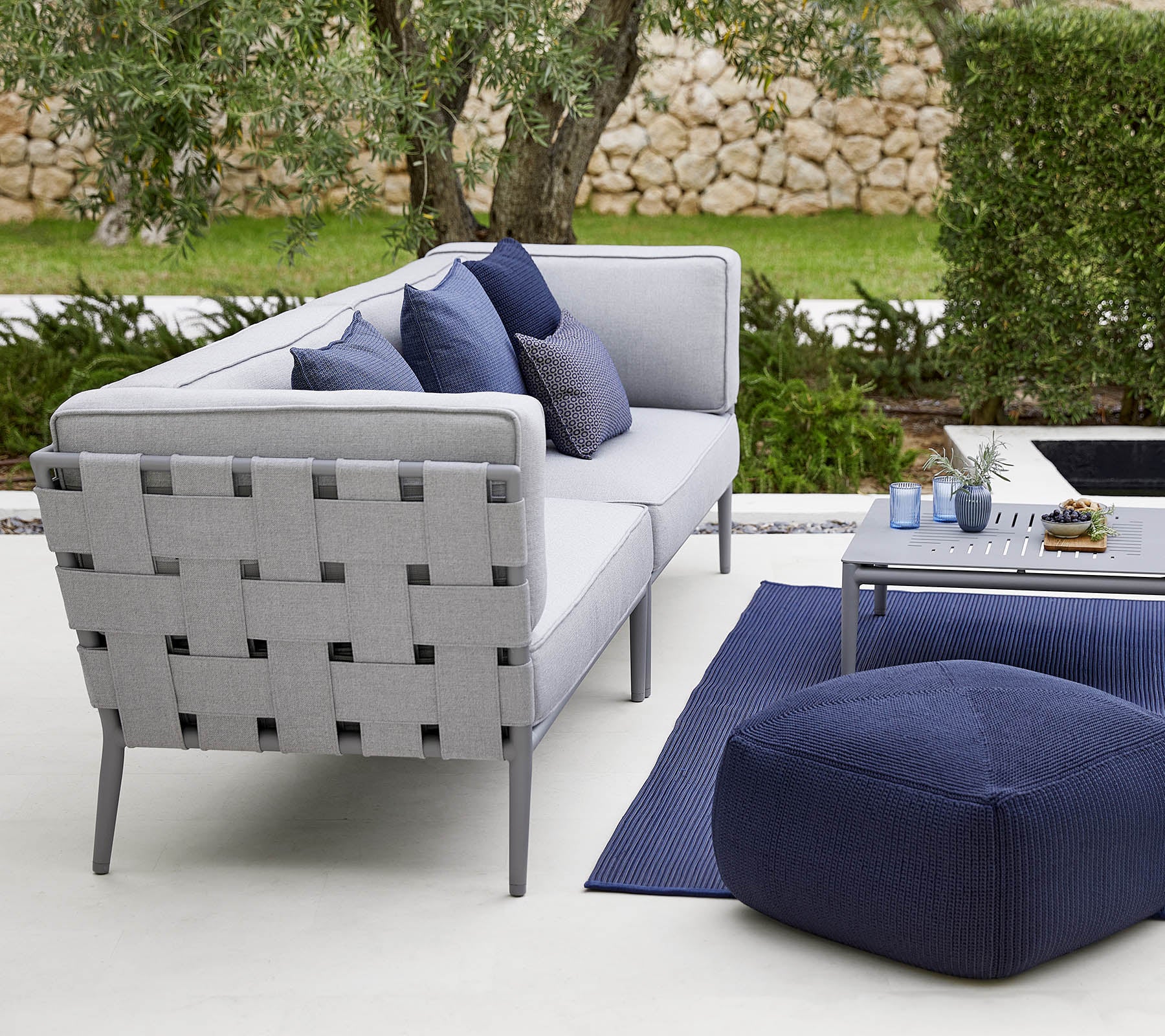 Disposición moderna de asientos al aire libre con un sofá gris con cojines decorativos, una mesa de café elegante y un pouf acogedor.
