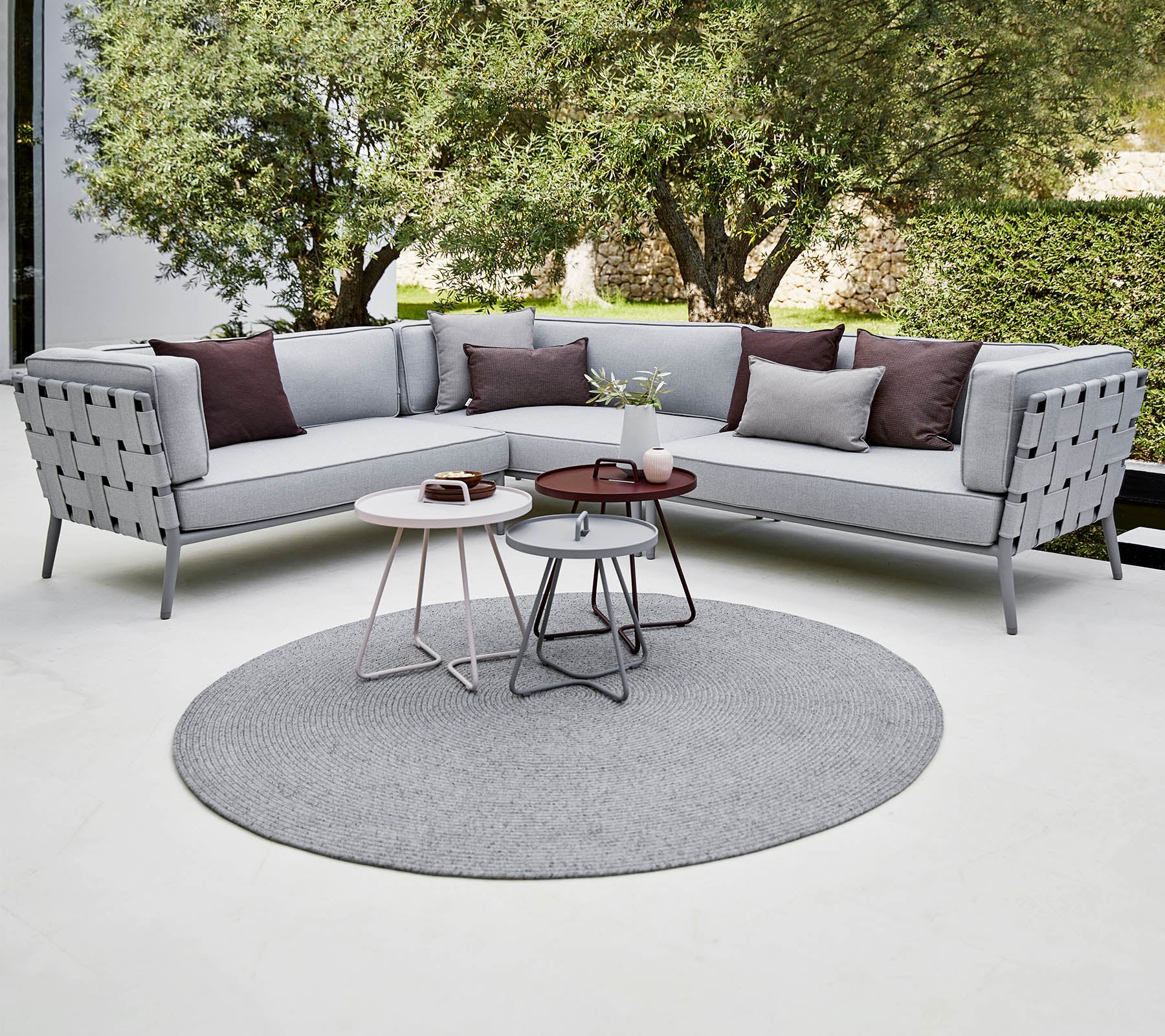 Sofá seccional moderno en gris claro con almohadas decorativas, acompañado de mesas de café elegantes sobre una alfombra redonda. Vegetación exuberante en el fondo.