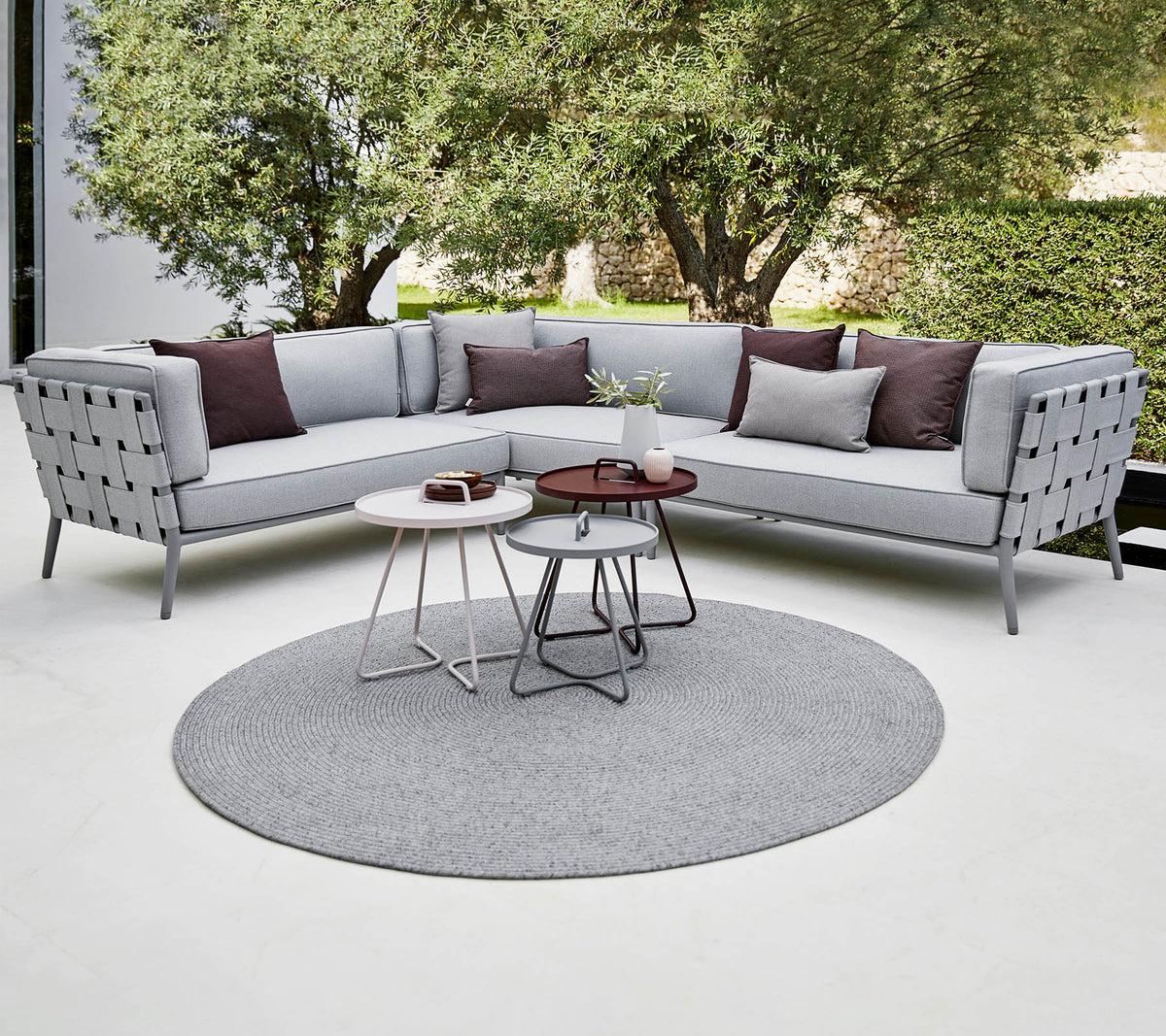 Sofá seccional moderno en gris claro con almohadas decorativas, acompañado de mesas de café elegantes sobre una alfombra redonda. Vegetación exuberante en el fondo.