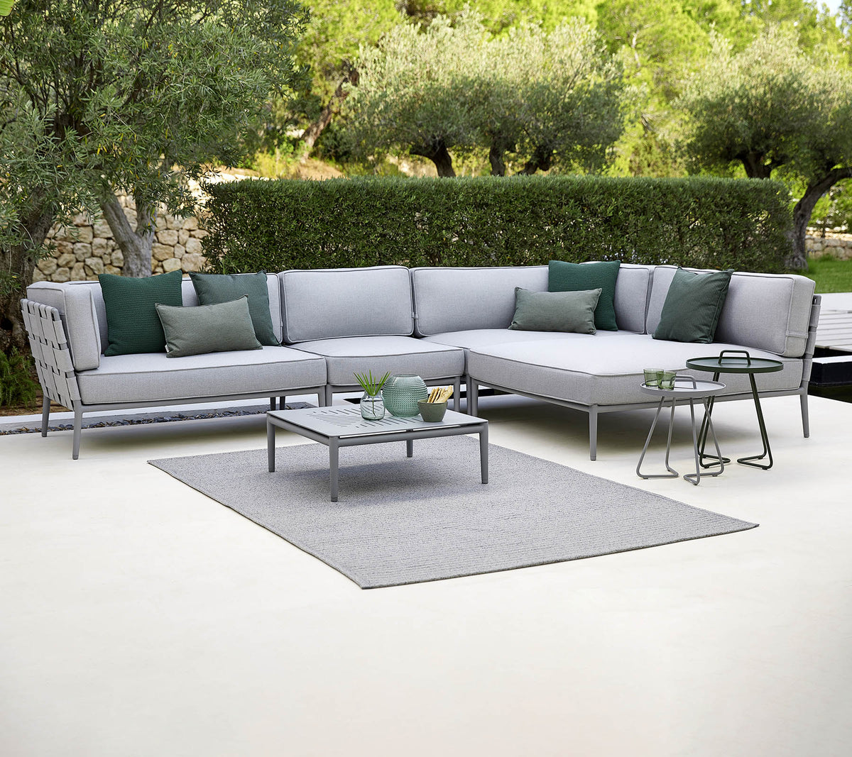 Sofá seccional exterior elegante con cojines verdes, una mesa de café y una alfombra, con un fondo de vegetación.