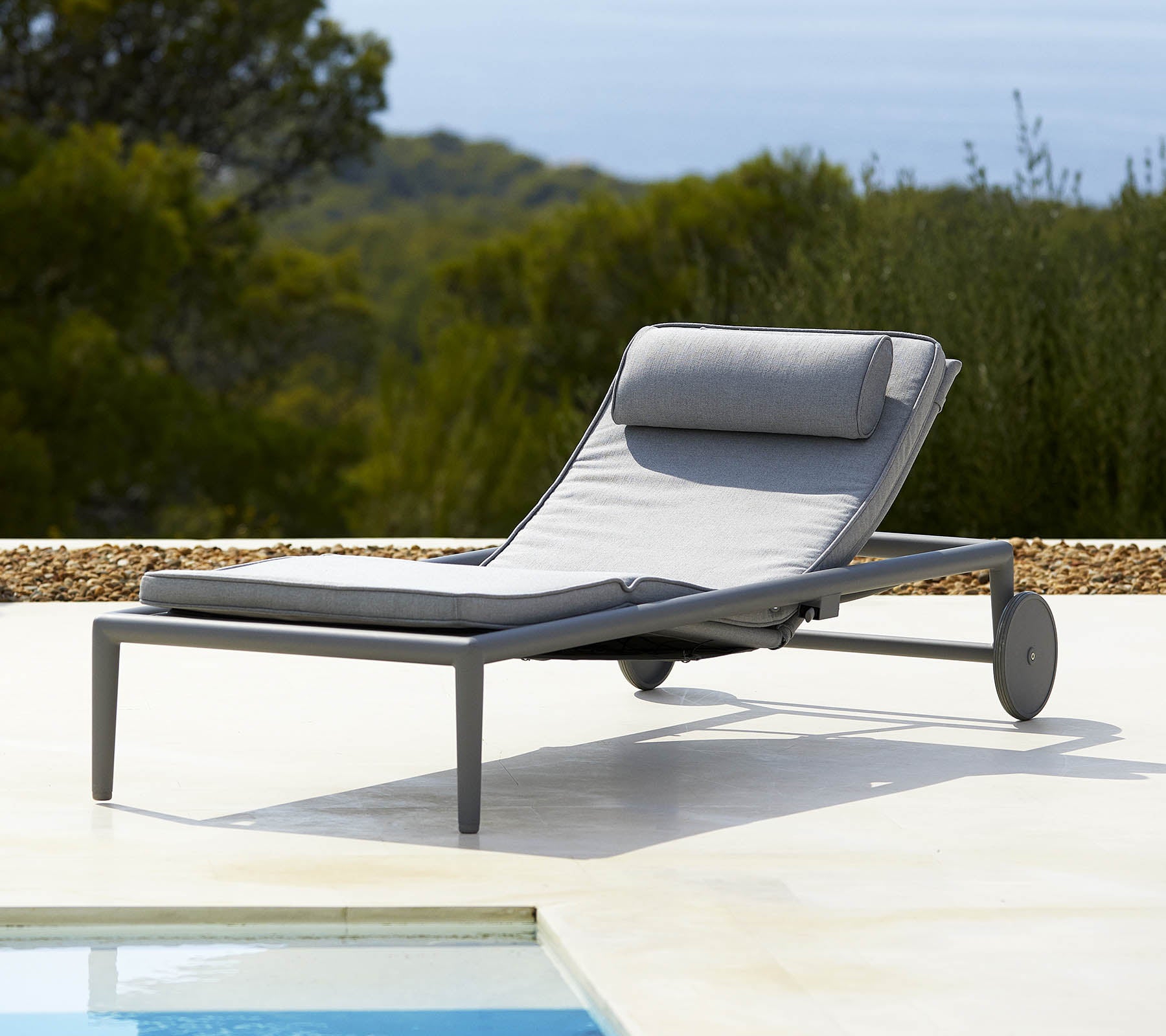Sillón lounge cómodo con un cojín gris y respaldo, con ruedas para fácil movilidad, al lado de una piscina y vegetación exuberante en el fondo.