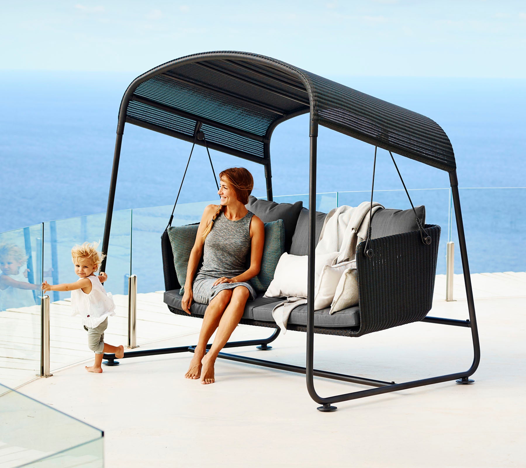Un sillón columpio elegante con cojines cómodos, que presenta a una mujer relajándose y a un niño jugando cerca, con un fondo oceánico sereno.
