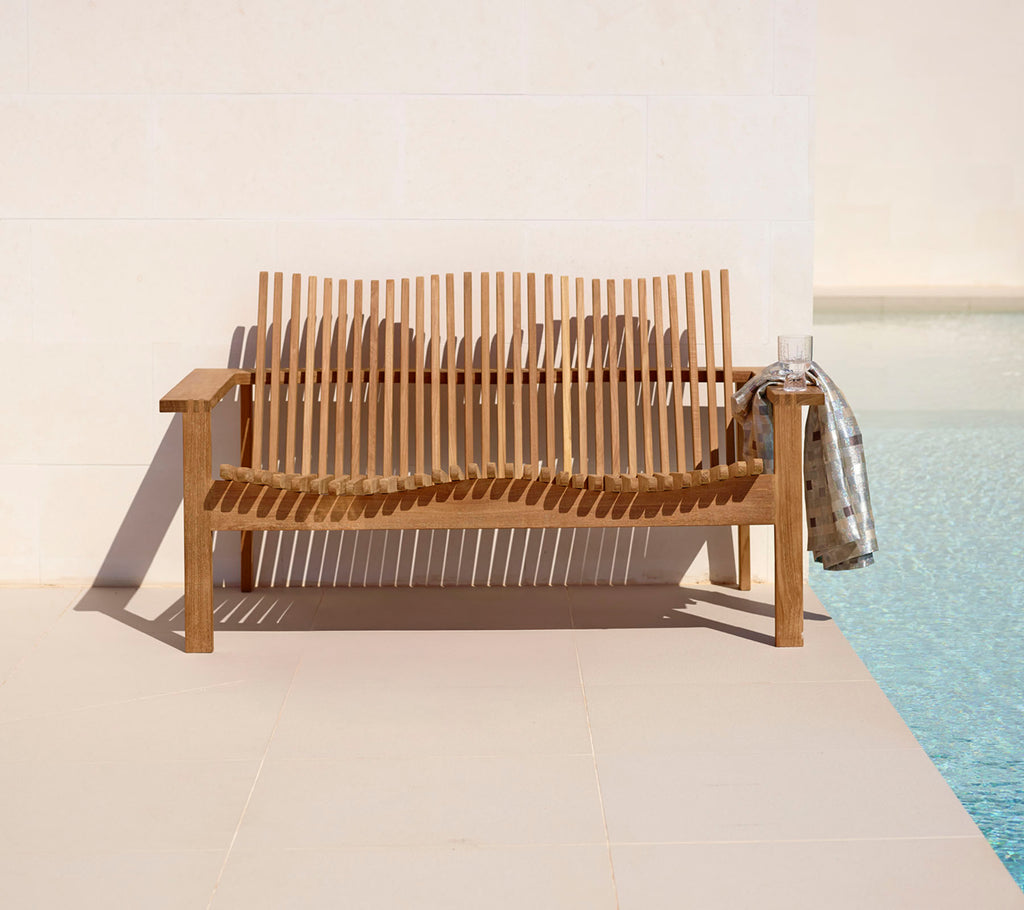 Sofá de madera elegante para dos personas con diseño de listones, complementado con una manta acogedora, junto a una piscina tranquila. Perfecto para relajarse.