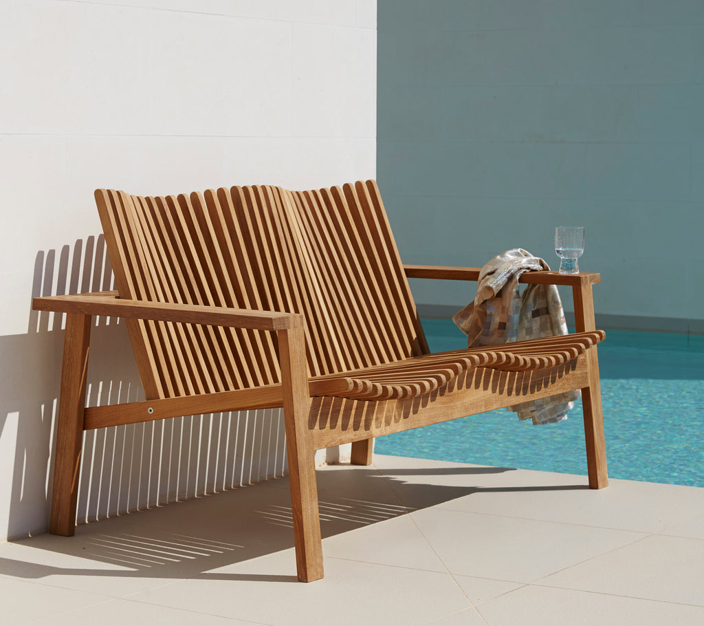 Vista lateral de un sofá de dos plazas de madera elegante con diseño de lamas, acompañado de una bebida y una toalla. Ideal para un asiento relajado.