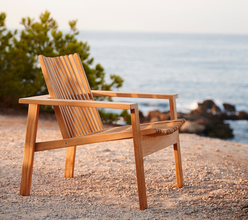 Vista lateral de una silla de lounge de madera con respaldo de lamas, situada cerca de una serena escena acuática y rodeada de vegetación.