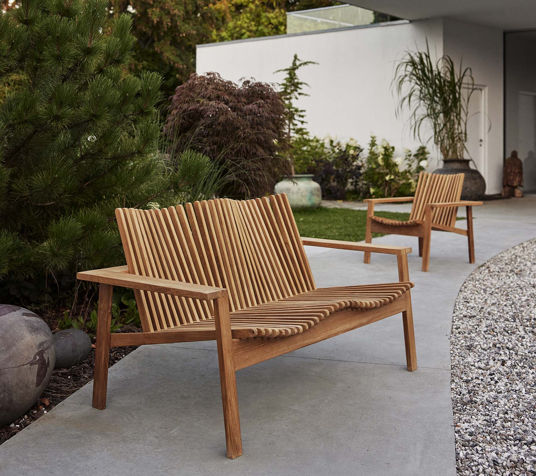 Dos sillones Amaze de madera y un sofá a juego presentan un diseño en listones, rodeados de vegetación y elementos decorativos.