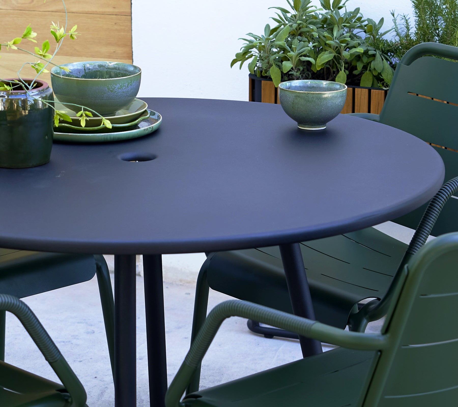 Mesa redonda gris con sillas verdes de Copenhague, con cuencos decorativos y vegetación de fondo.