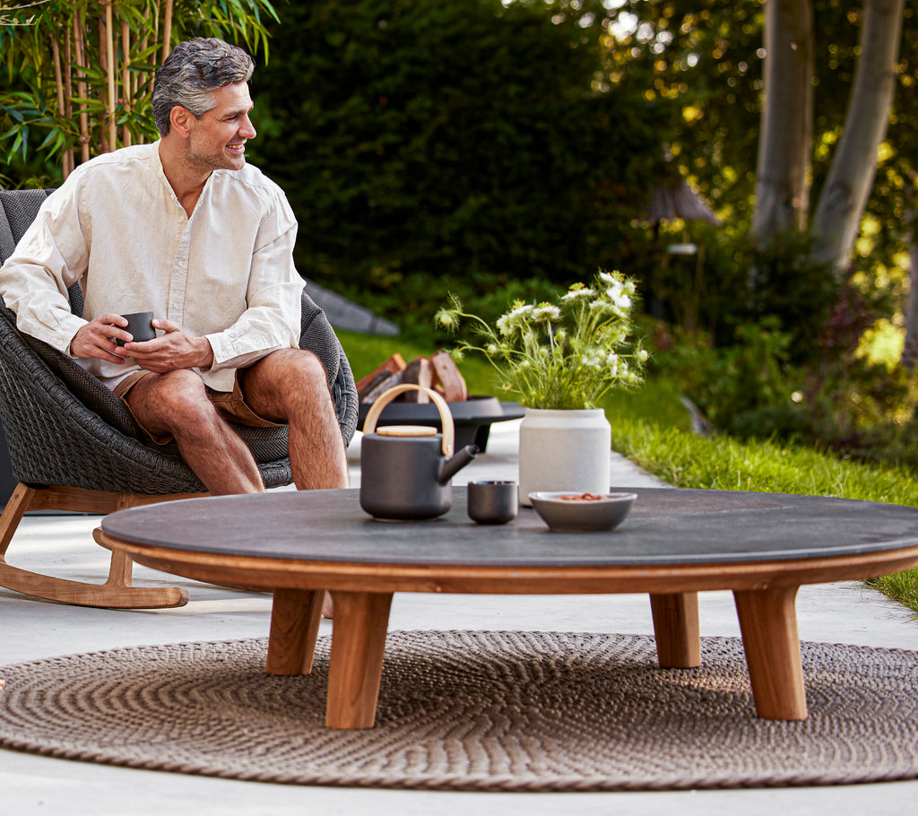 Un hombre disfruta de una bebida mientras está sentado en una silla mecedora junto a una mesa de café redonda adornada con vajilla elegante y vegetación.