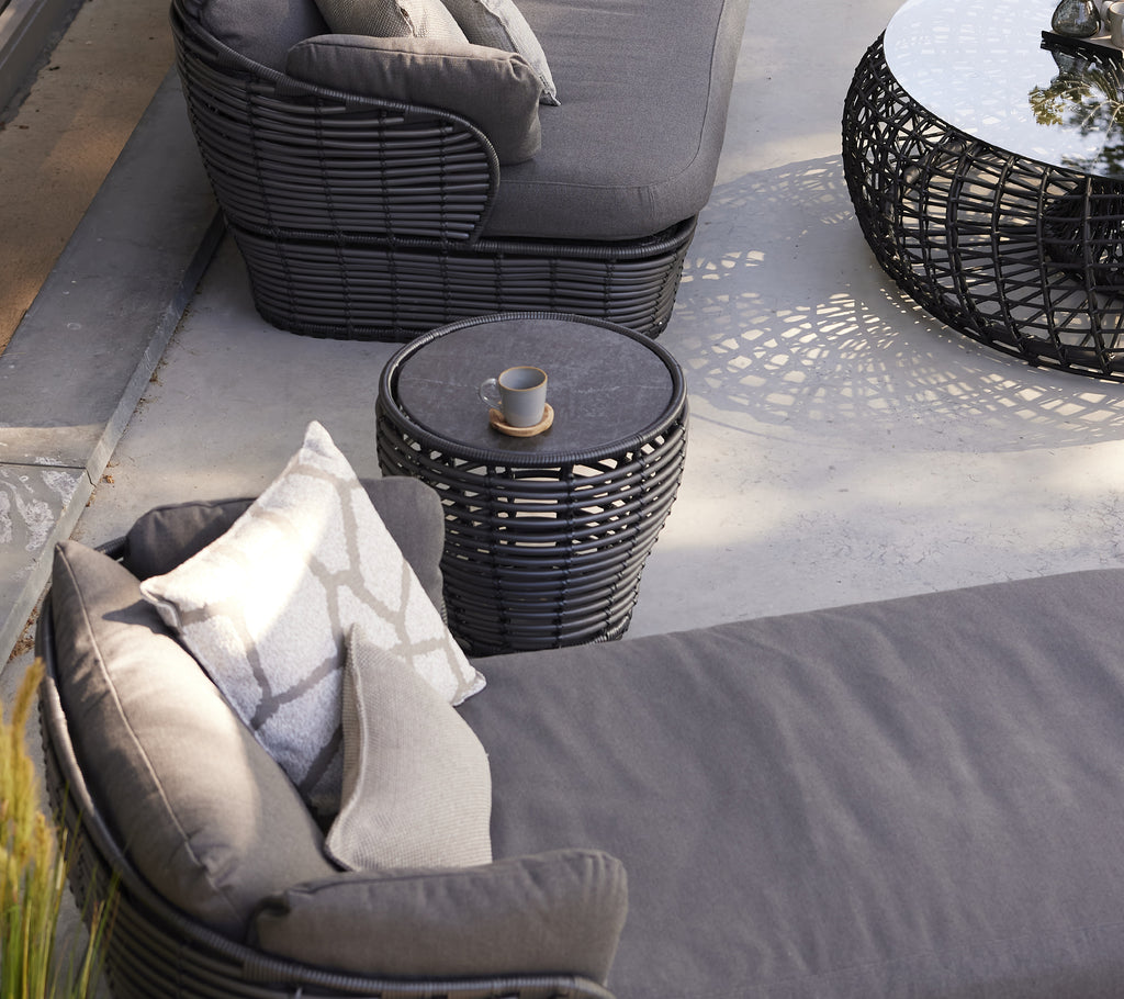 Una pequeña mesa de café de grafito en forma de cesta al lado de una silla moderna con cojines, mejorando un área de estar elegante.