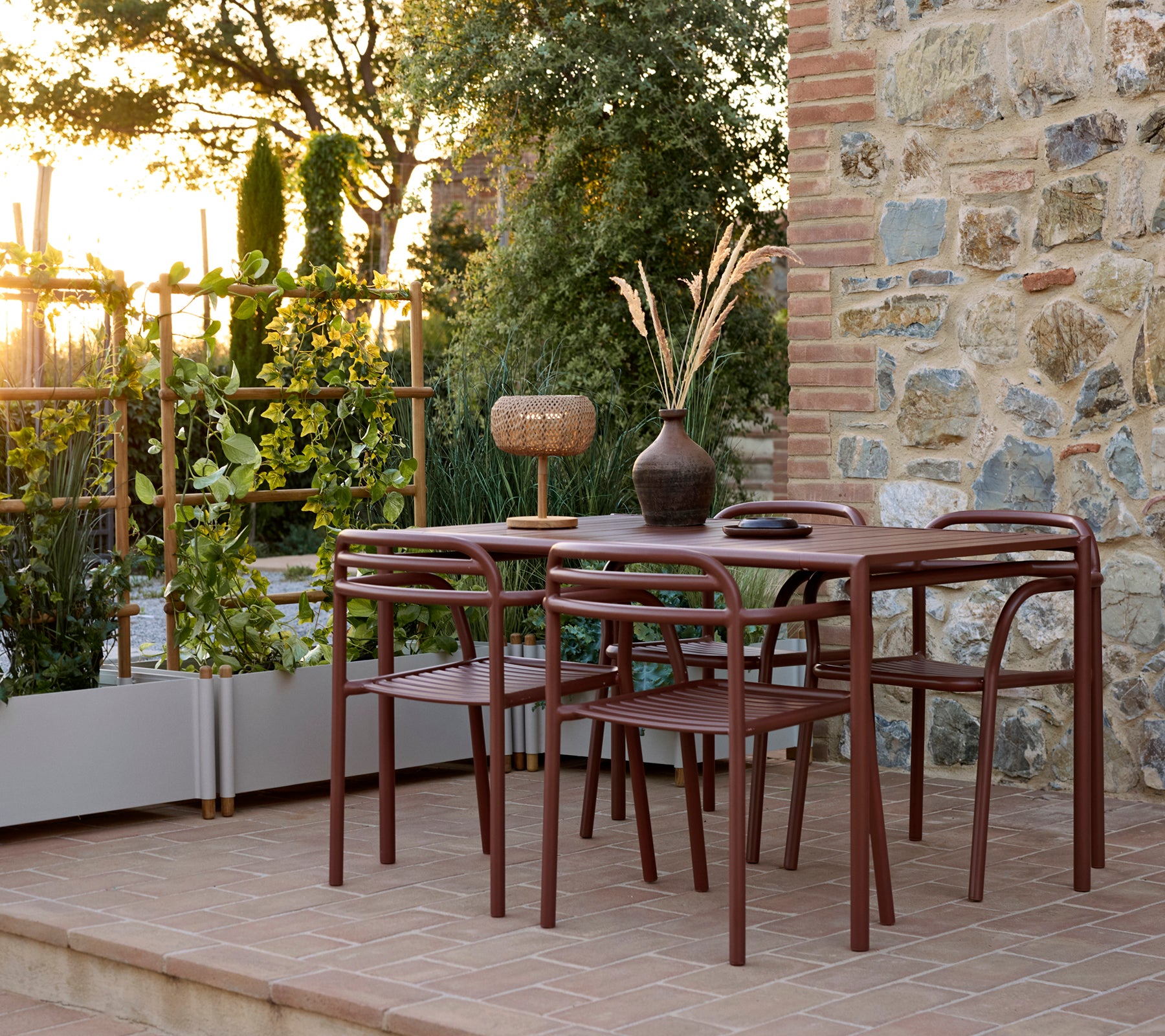 Configuración al aire libre con una mesa Bliss con sillas, una sillón Bliss y una maceta Sticks rodeada de vegetación al atardecer.