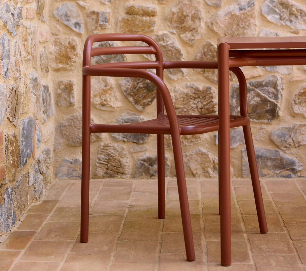 Sillón Bliss rojo desierto al lado de una mesa a juego, colocado contra una pared de piedra texturizada. Un arreglo de asientos elegante y moderno.