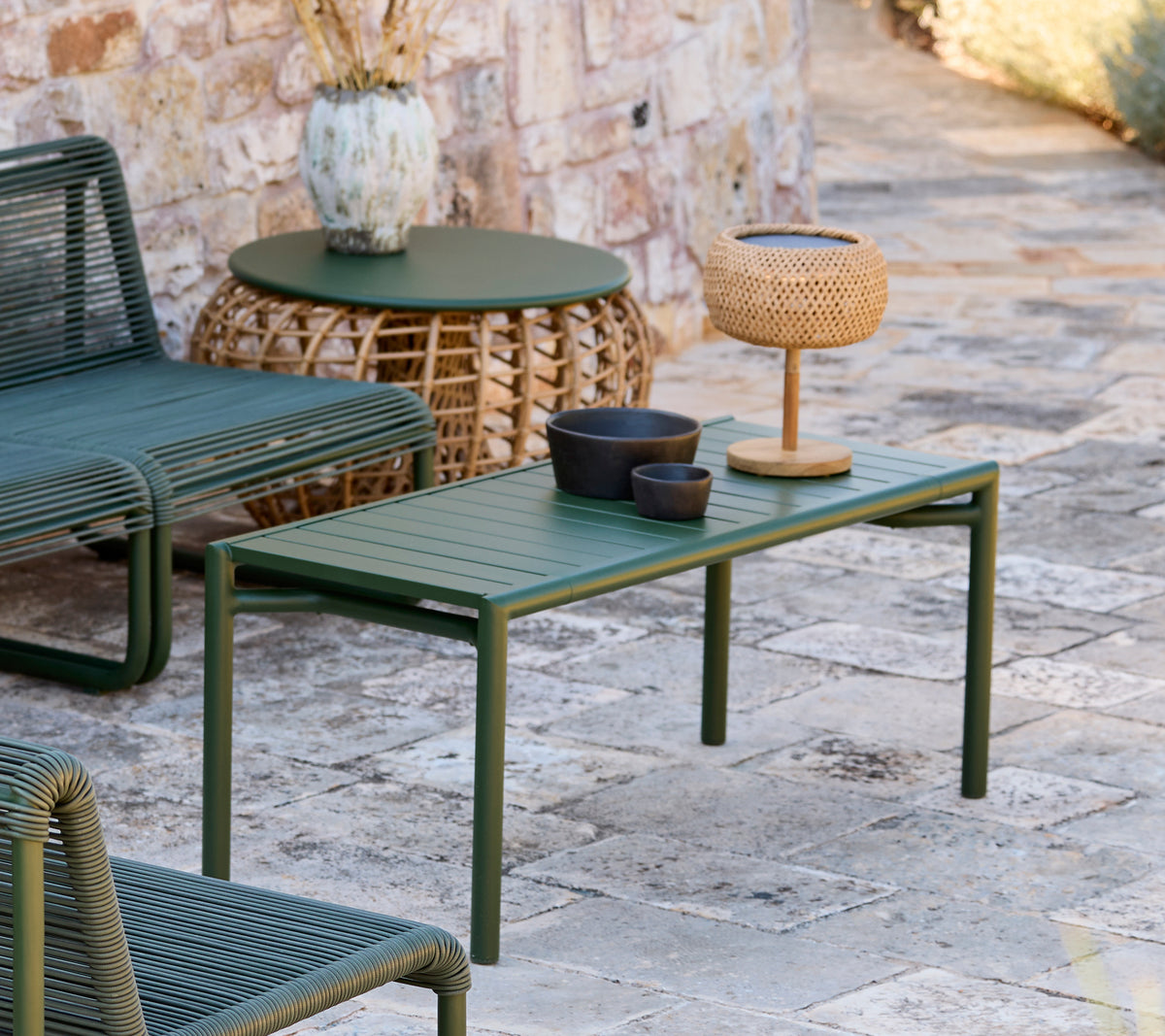 Mesa verde con cuenco negro, jarrón marrón y lámpara tejida en un área de salón moderno al aire libre.