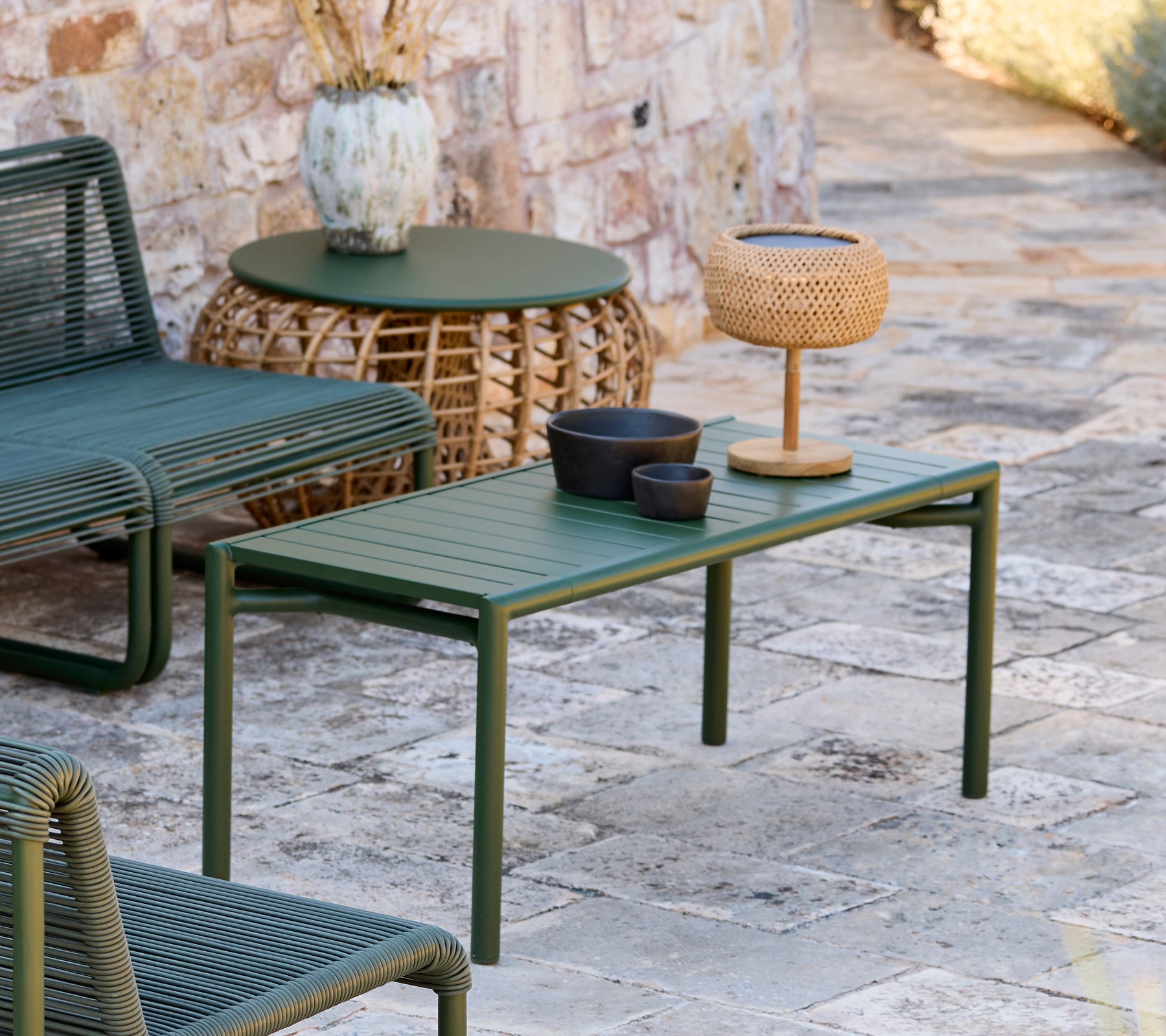 Mesa verde con cuenco negro, jarrón marrón y lámpara tejida en un área de salón moderno al aire libre.