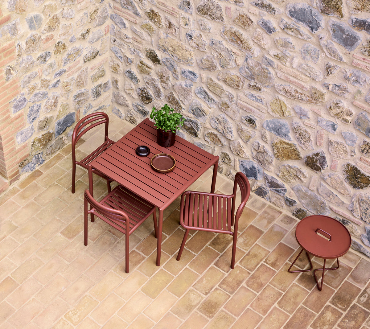 Una mesa y sillas en un tono rojo desértico, junto a una pequeña mesa redonda, contra una pared de piedra texturizada con una planta en maceta.