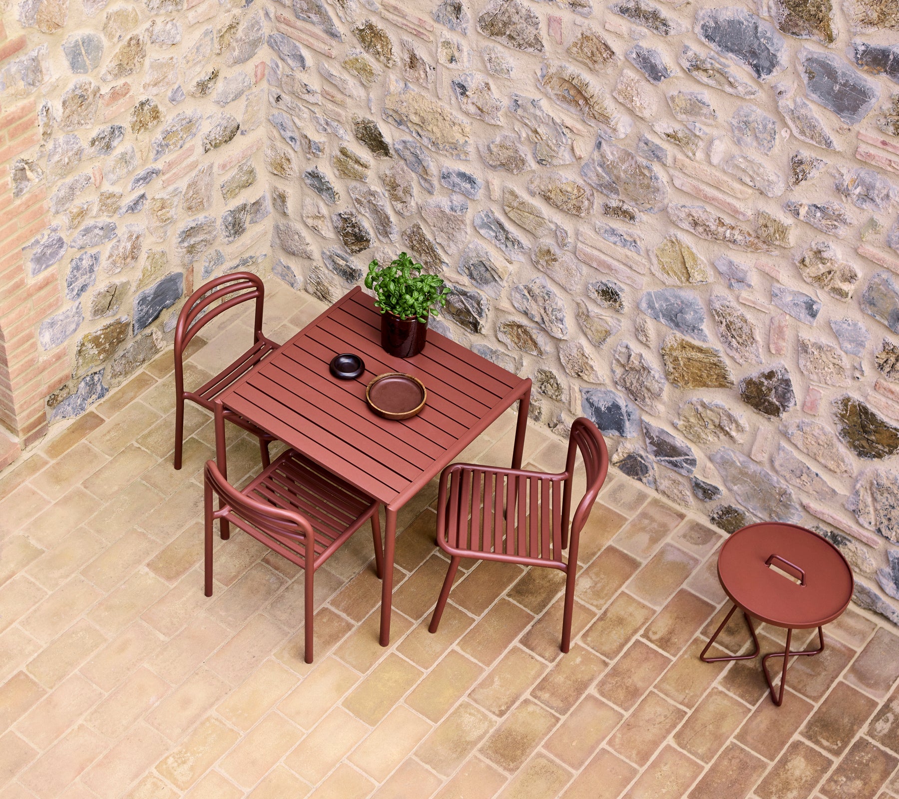 Una mesa y sillas en un tono rojo desértico, junto a una pequeña mesa redonda, contra una pared de piedra texturizada con una planta en maceta.
