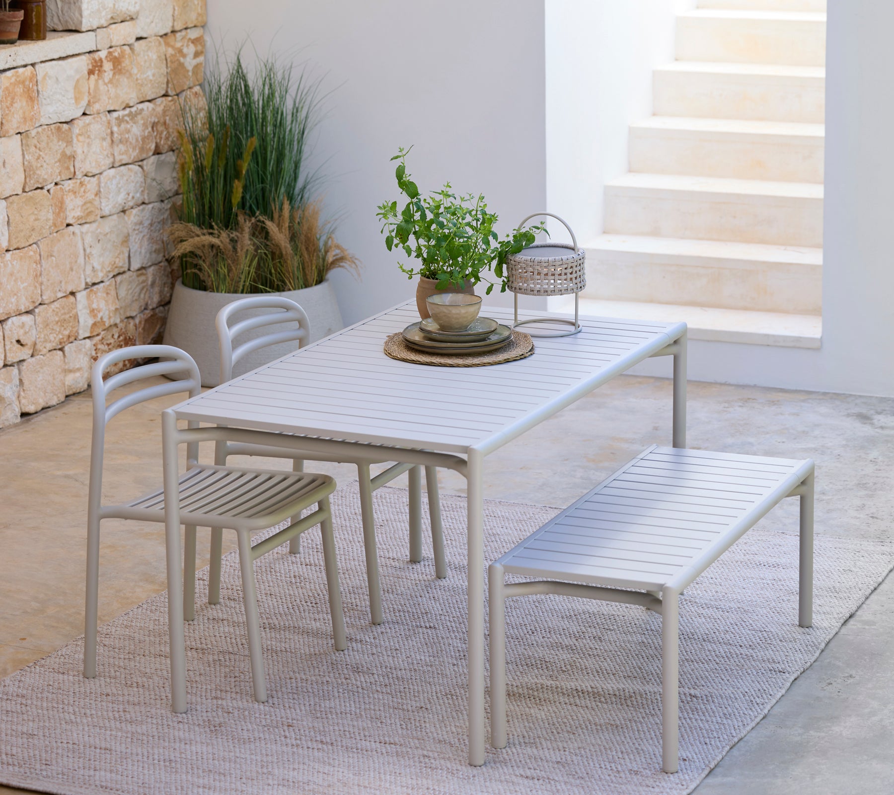 Conjunto de comedor gris claro que incluye una mesa, sillas y un banco en un entorno exterior moderno.