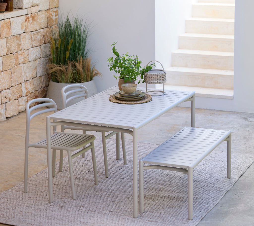 Conjunto de comedor gris claro que incluye una mesa, sillas y un banco en un entorno exterior moderno.