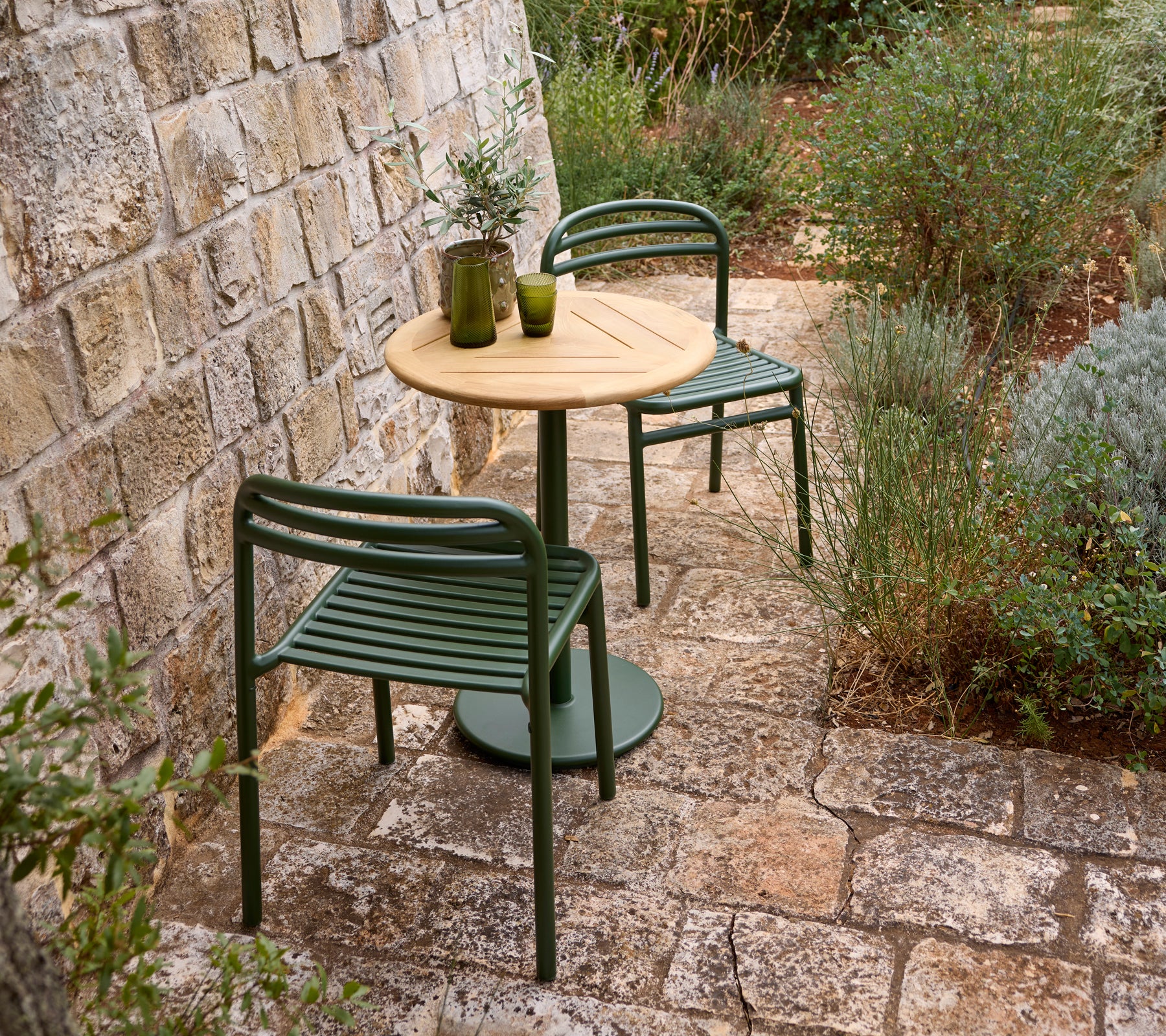 Configuración al aire libre con una silla verde, una mesa de madera y un jarrón de vidrio.