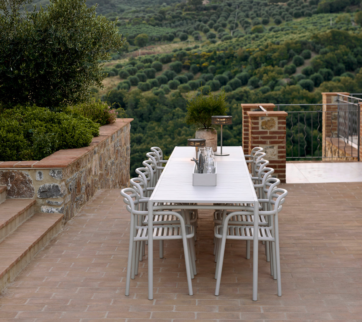 Una disposición de asientos elegante con sillas y mesa Bliss en color arena, complementada por una lámpara Illusion, en un paisaje exuberante.