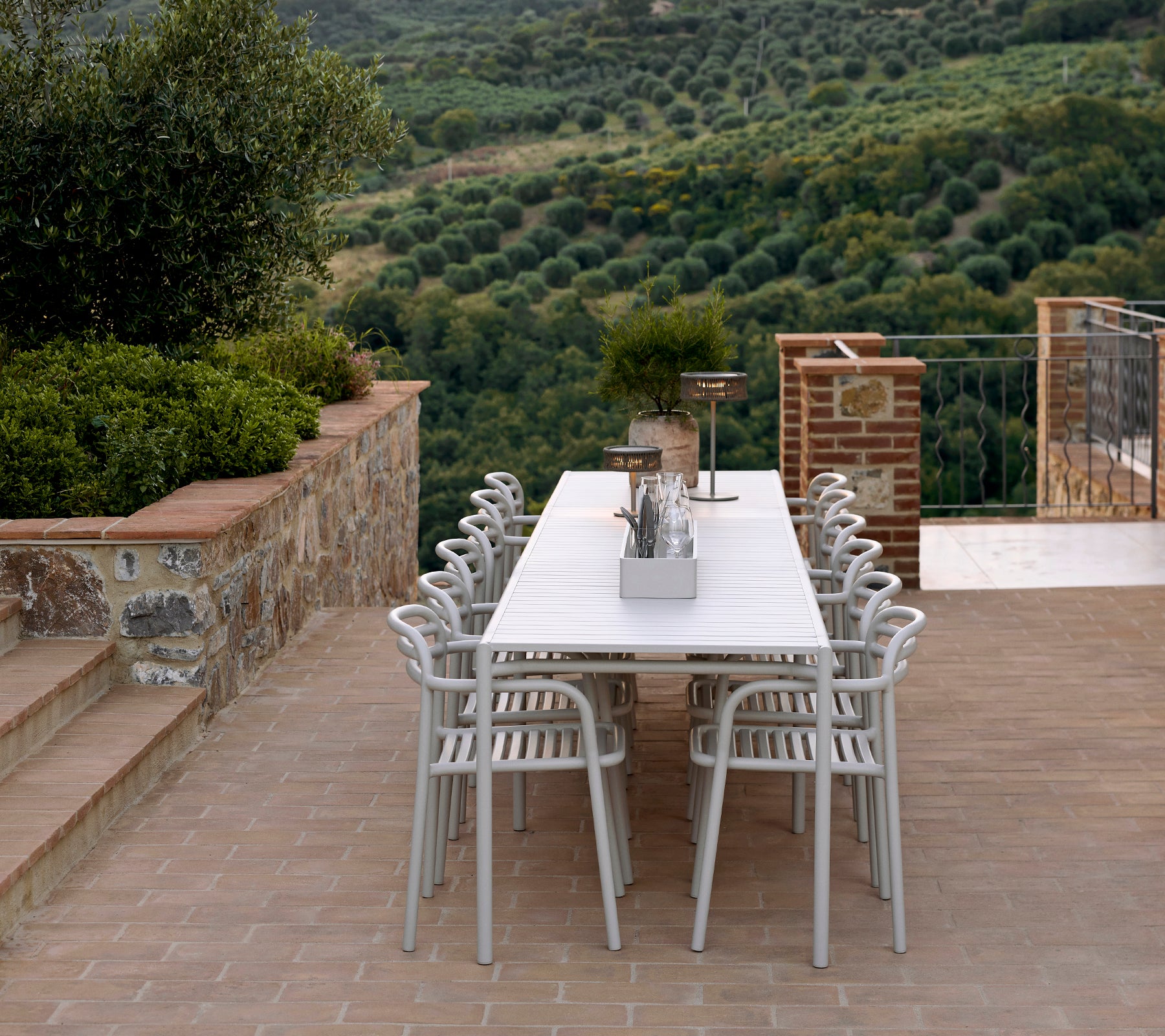 Una disposición de asientos elegante con sillas y mesa Bliss en color arena, complementada por una lámpara Illusion, en un paisaje exuberante.