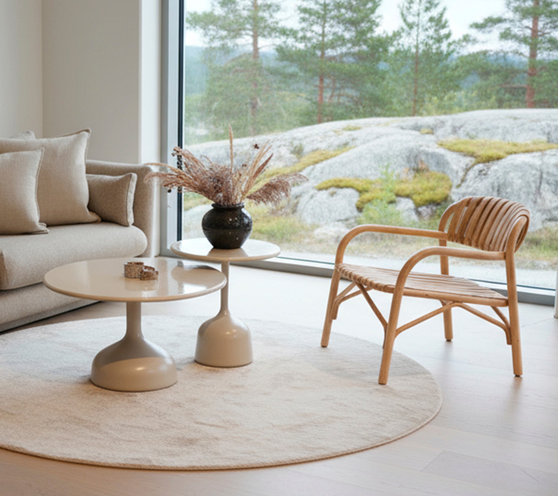 Salón moderno con un sofá, mesa redonda, jarrón negro y silla de ratán cerca de una gran ventana.
