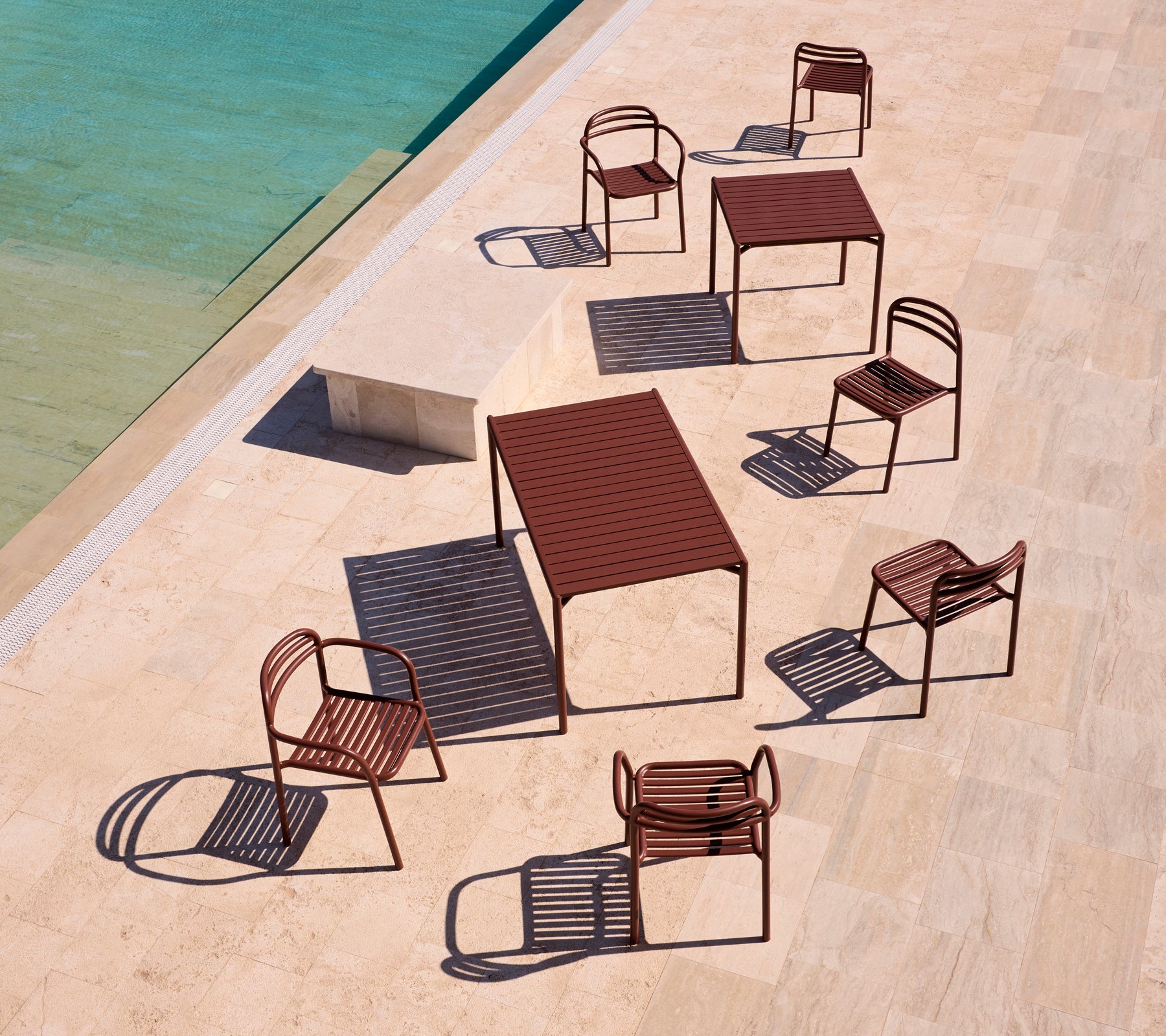 Muebles de exterior de la serie Bliss en rojo desierto, dispuestos alrededor de una piscina en una terraza iluminada por el sol, proyectando sombras juguetonas.