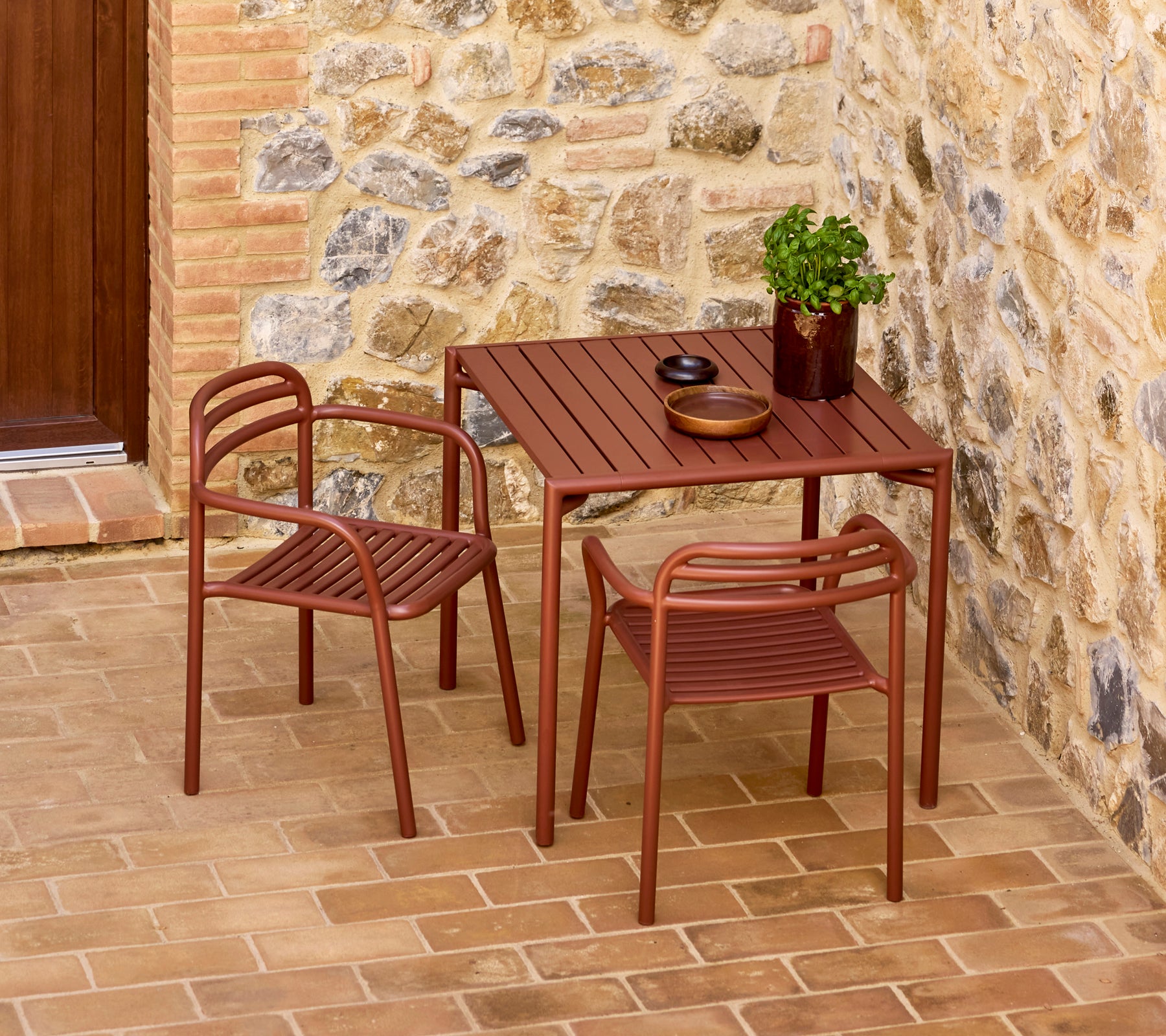 Mesa Bliss rojo desértico elegante con dos sillas a juego, acompañada de una planta decorativa y vajilla simple contra un fondo de piedra.