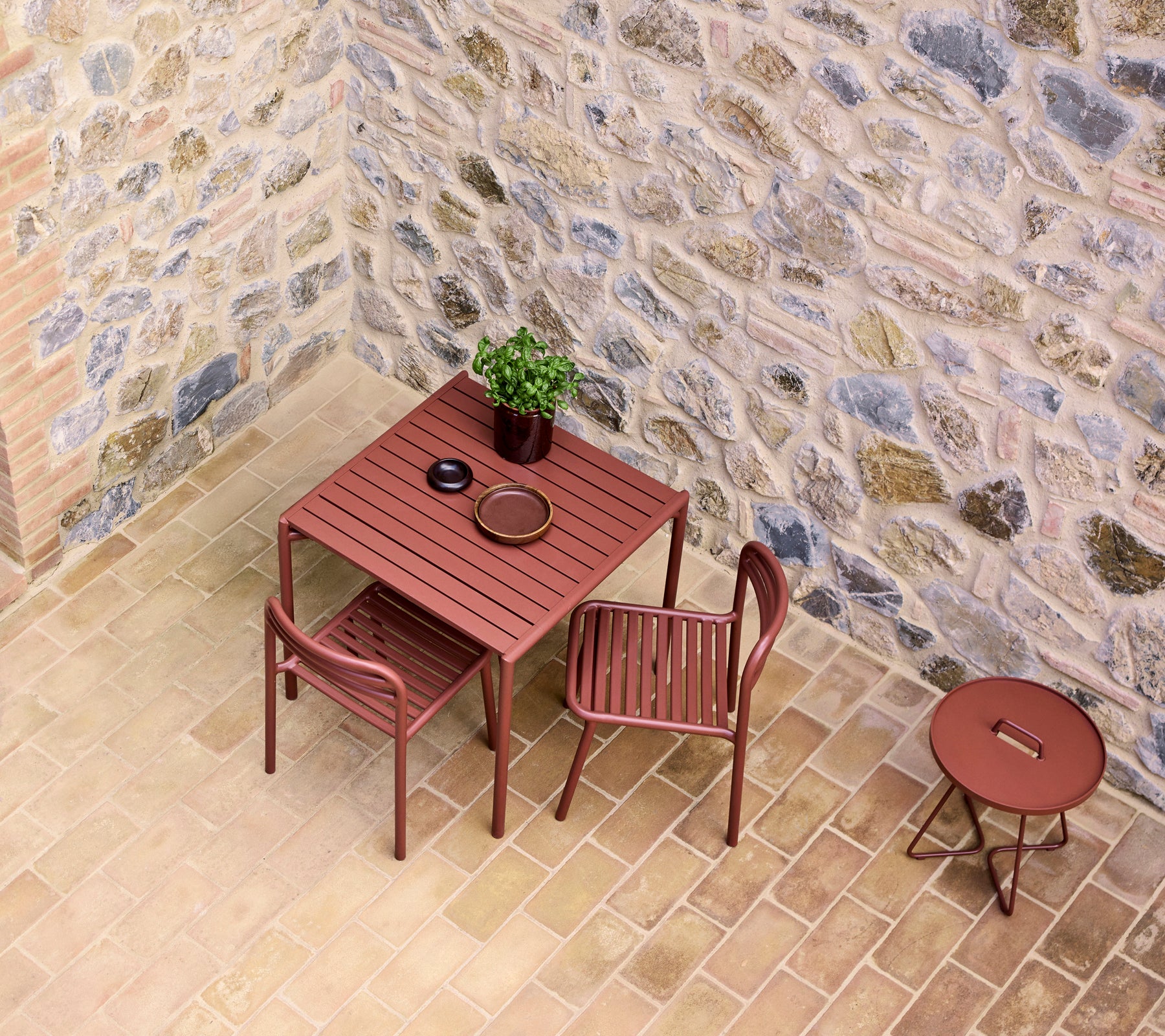 una mesa de café roja con dos sillas, con una pequeña planta y un plato, contra un fondo de pared de piedra.