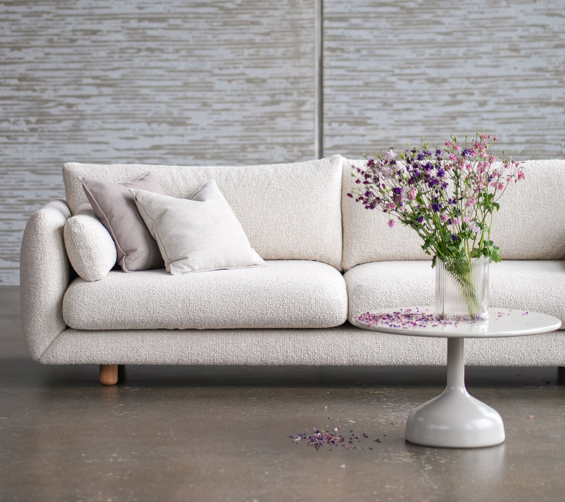 Sofá de color claro con patas de teca, adornado con cojines, complementado por una mesa redonda y un jarrón con flores frescas.