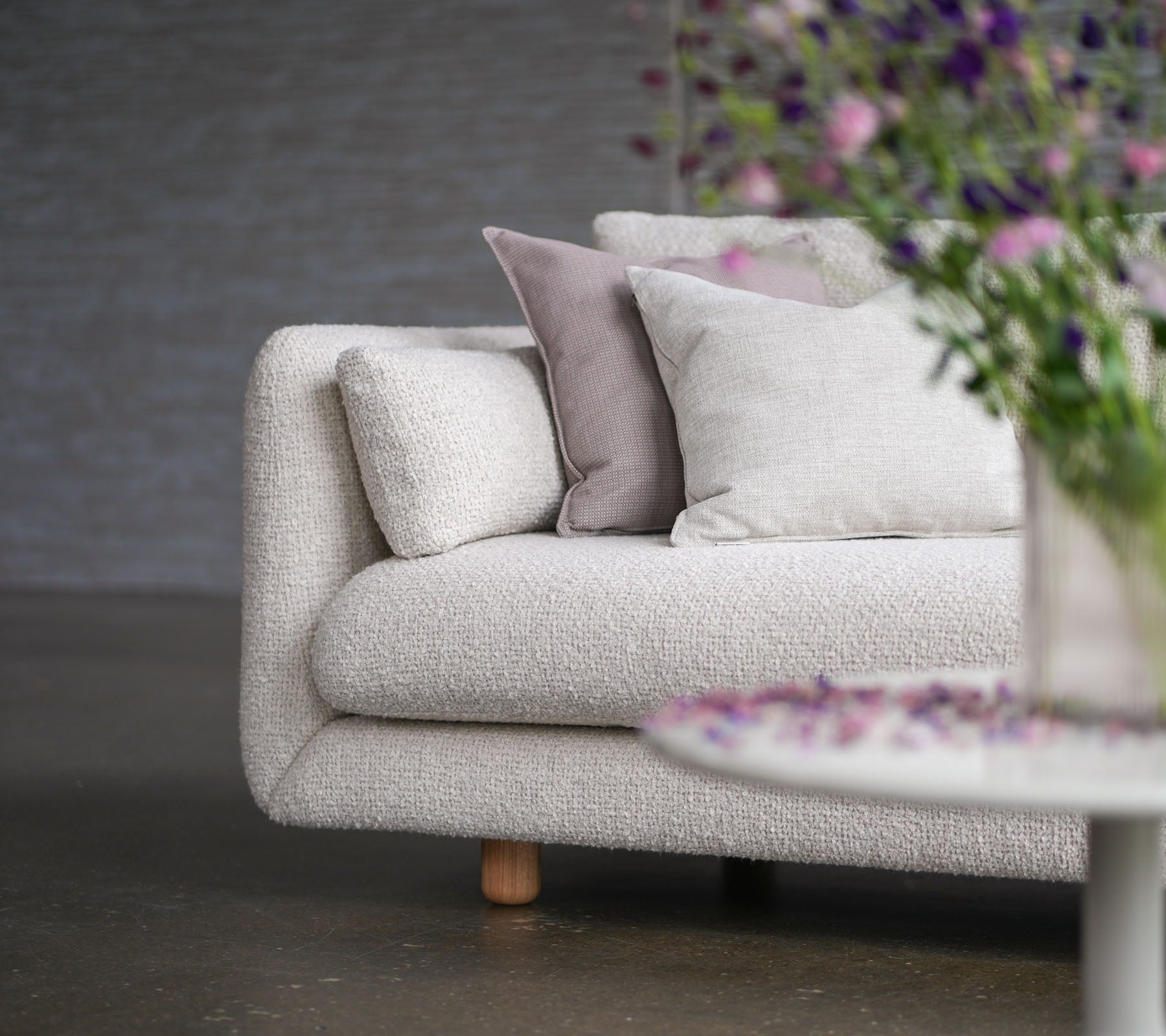 Sofá suave con tapicería color arena, cojines decorativos, patas de teca y un jarrón de flores, mostrando el estilo y la elegancia de Cane-line.