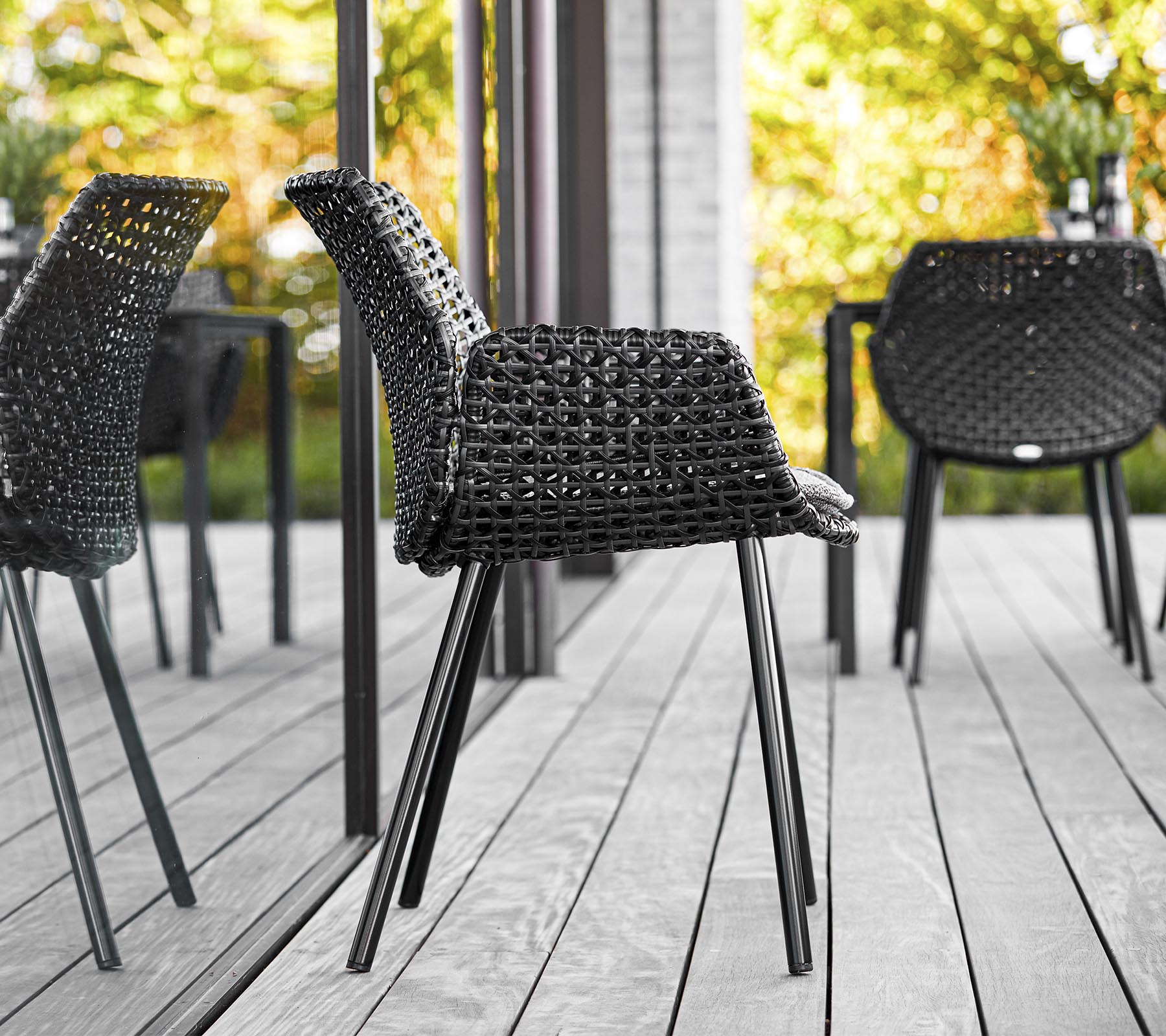 Silla trenzada negra elegante con patas esbeltas, situada contra un fondo de vegetación y reflejos, realzando la estética moderna al aire libre.