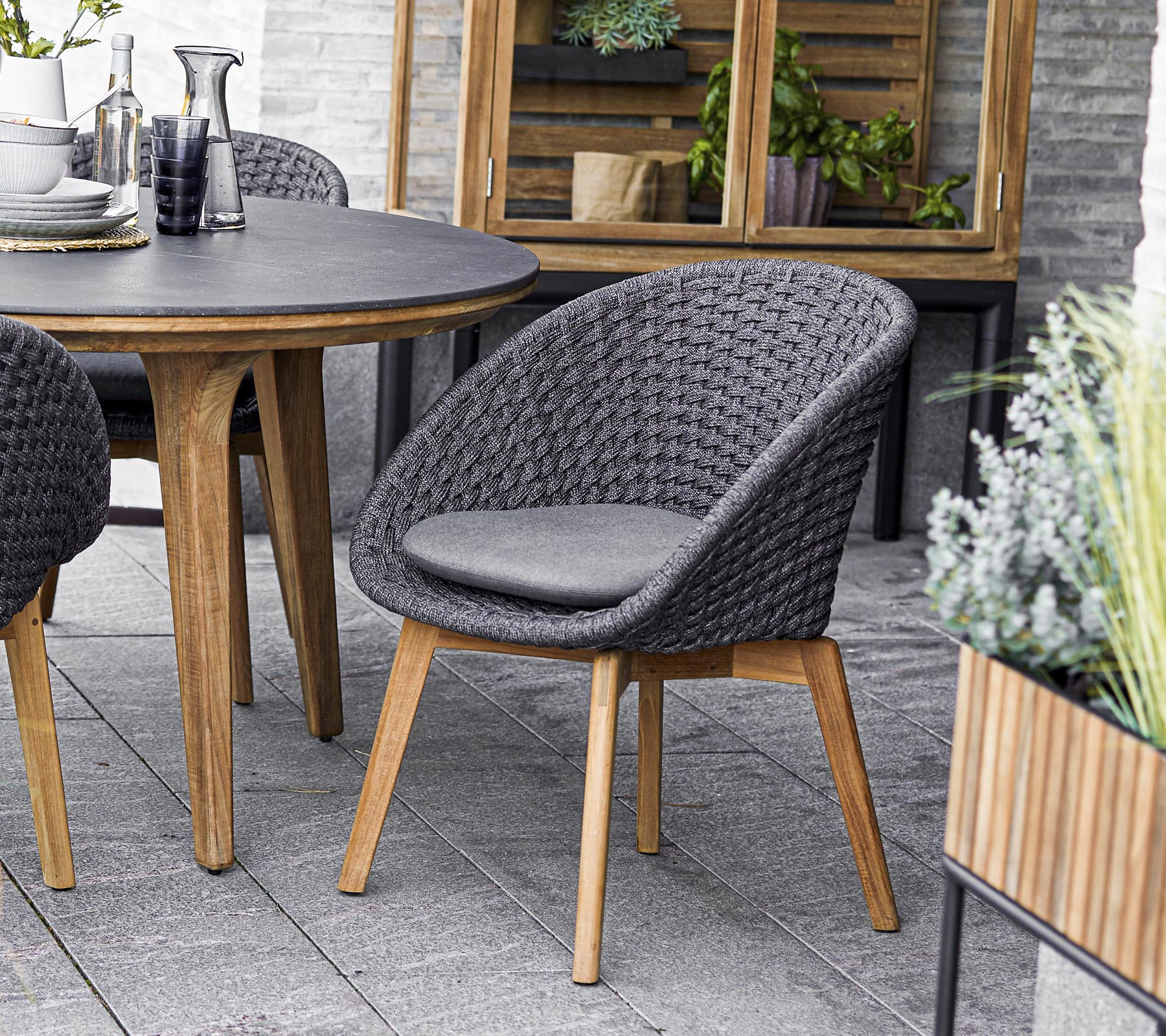 Silla tejida elegante con un asiento acolchado, acompañada de una mesa redonda y rodeada de vegetación en jardineras. Perfecto para reuniones sociales.