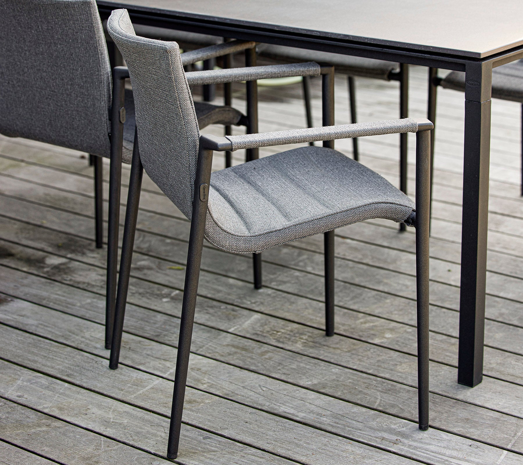 Silla gris elegante con reposabrazos ubicada al lado de una mesa negra elegante sobre una superficie de madera. Ideal para entornos exteriores modernos.