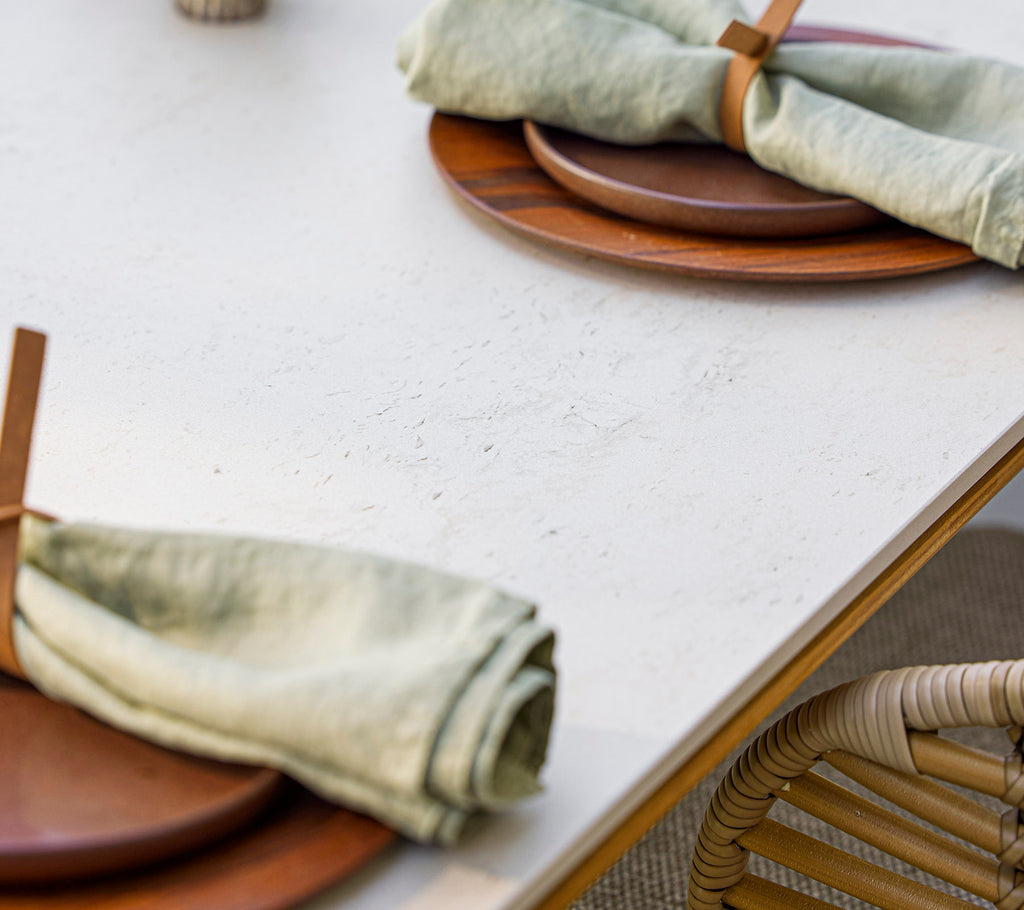 Mesa puesta con platos de madera y servilletas verdes elegantemente enrolladas aseguradas con un soporte de madera, creando una atmósfera natural y acogedora.