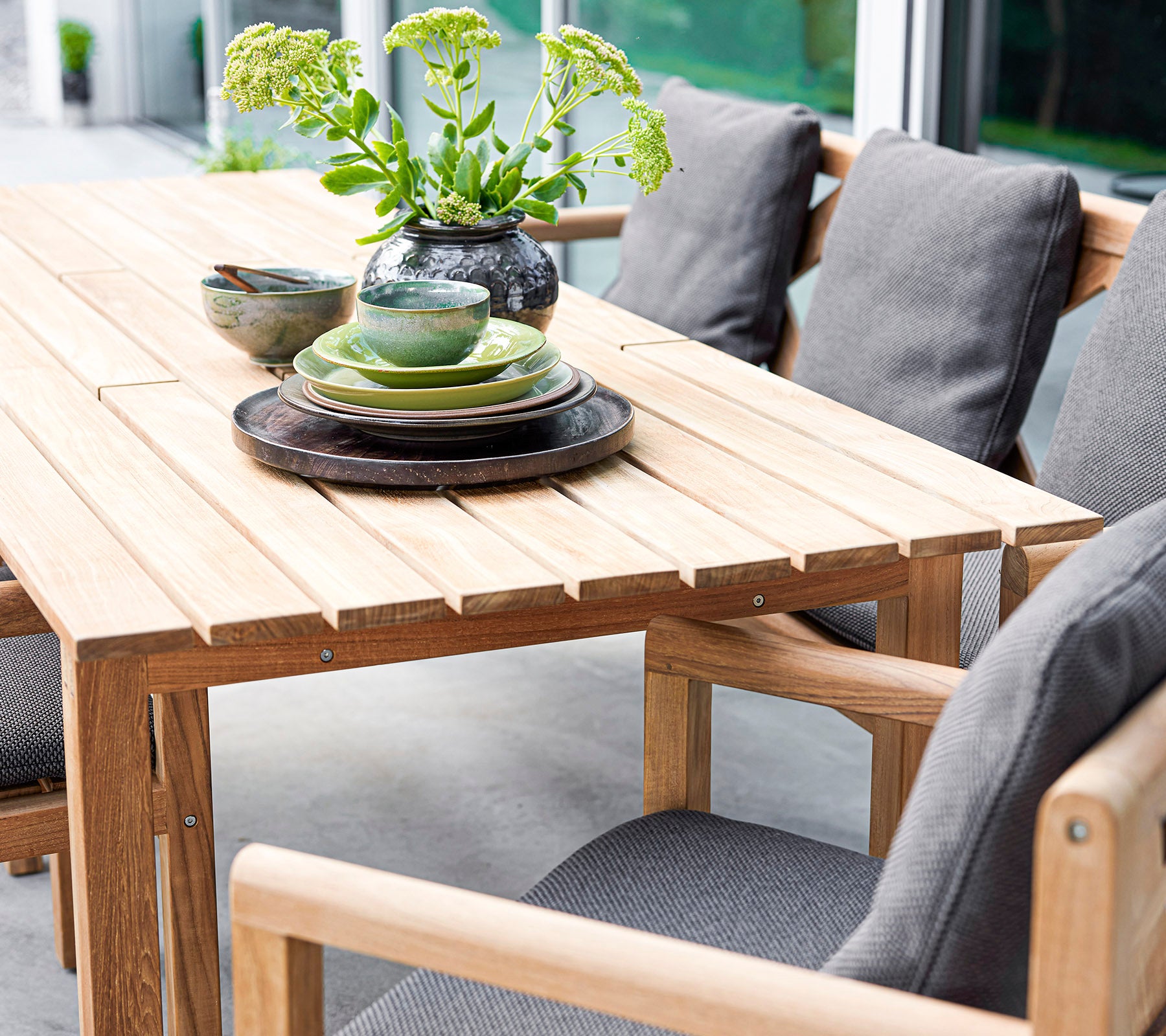 Juego de mesa de madera elegante con cojines, platos y cuencos, acentuado por un jarrón con vegetación. Perfecto para reuniones acogedoras.