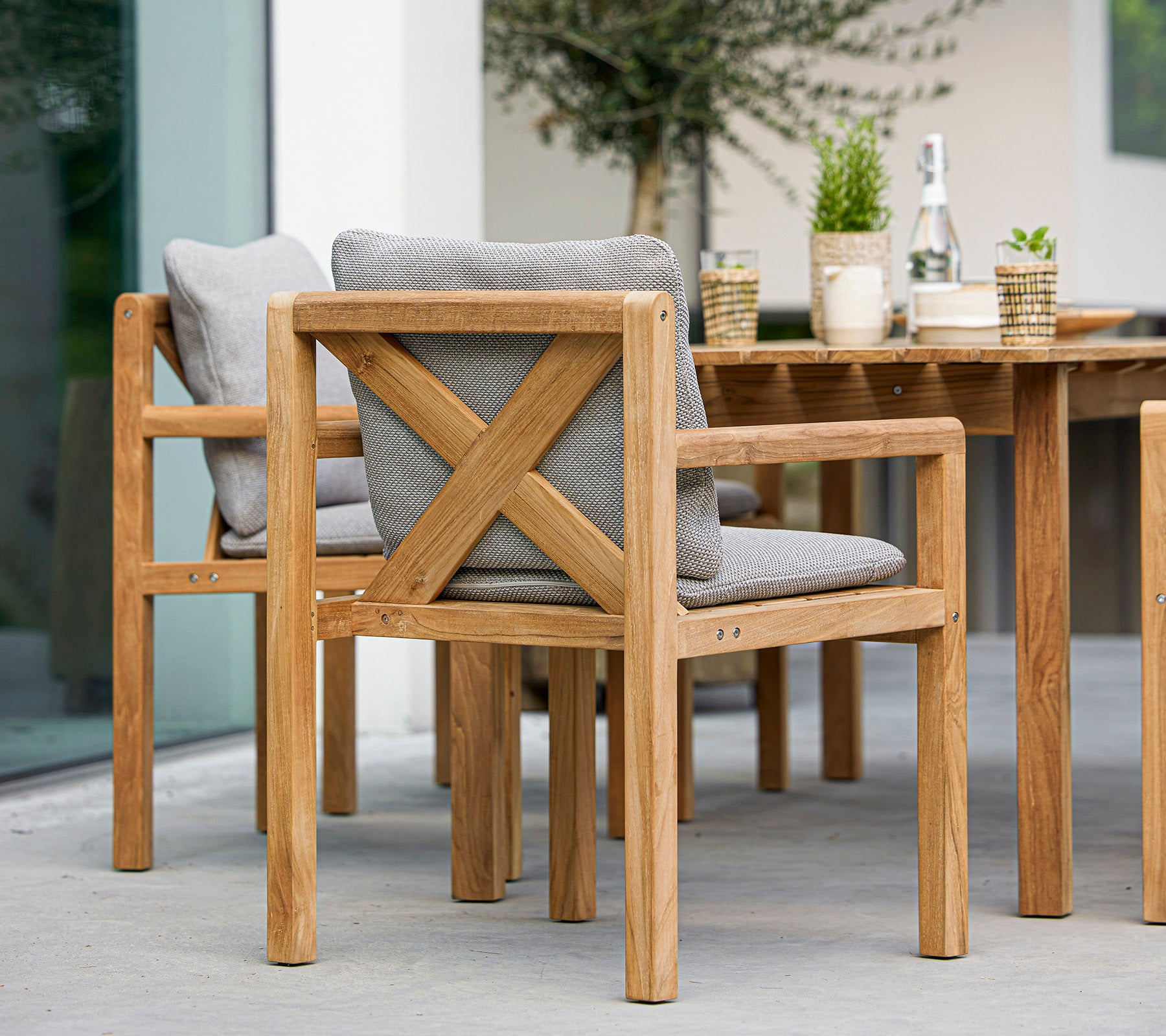 Sillas de madera con cojines grises y una mesa a juego, acompañadas de plantas decorativas y una botella, ideales para cenar al aire libre.