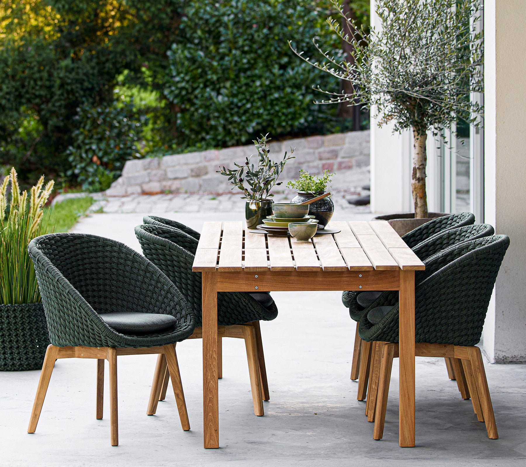 Una mesa de madera rodeada de sillas verdes elegantes, con plantas decorativas y un fondo sereno. Ideal para cenar o relajarse.