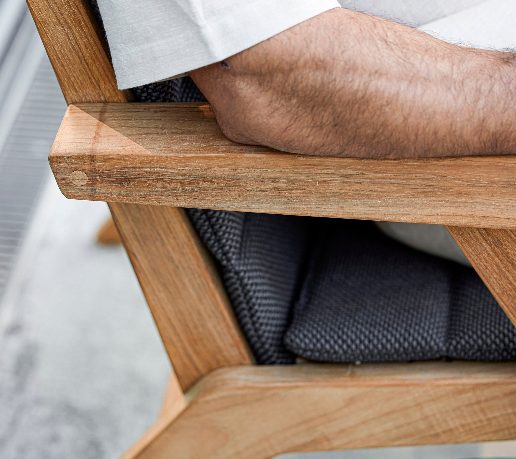 Un primer plano de un reposabrazos de silla de madera con un asiento acolchado, mostrando un brazo descansando sobre él, que exhibe una mezcla de materiales naturales y texturizados.