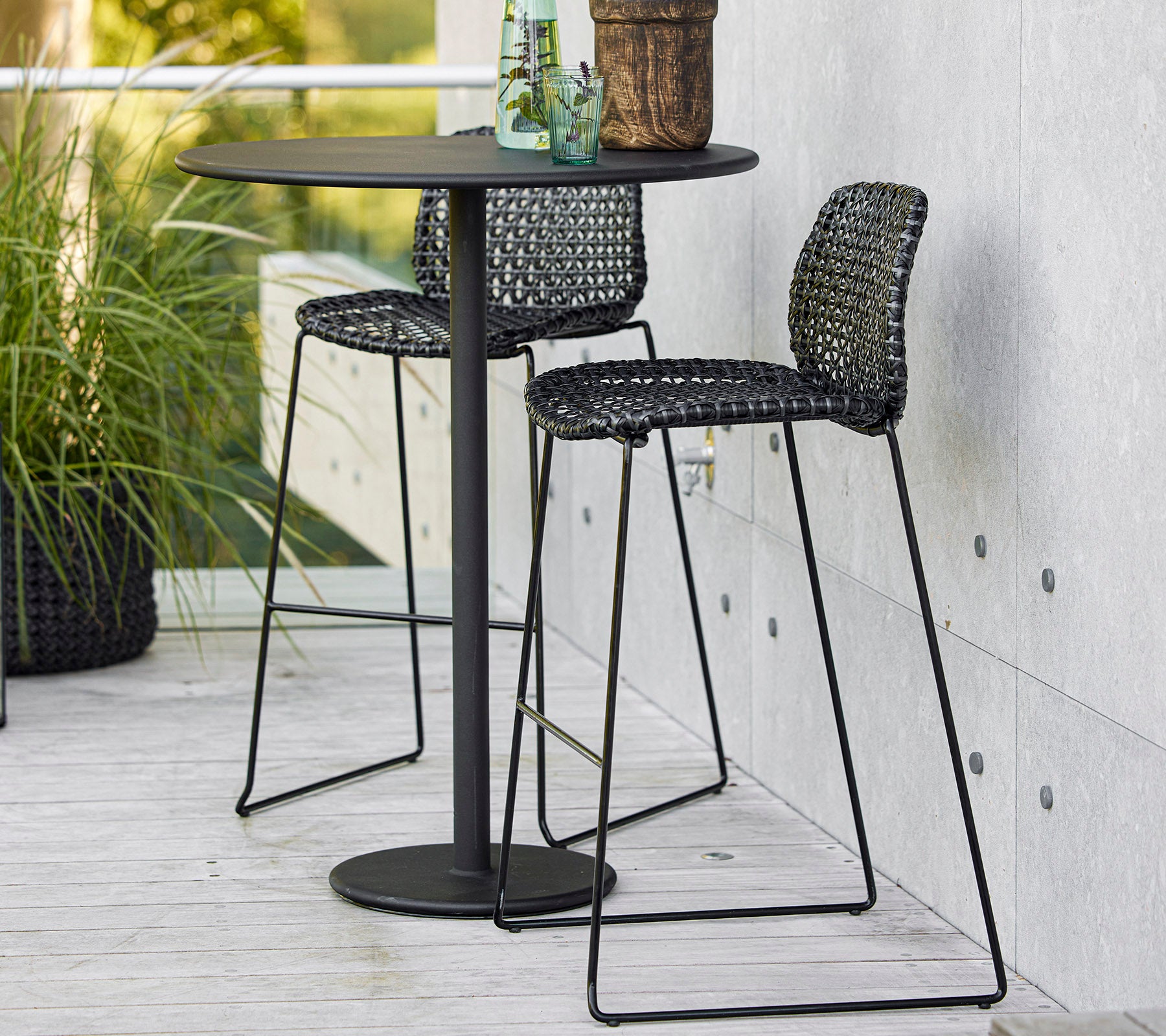 Mesa de bar negra elegante con dos sillas trenzadas y una planta en maceta, perfecta para una comida informal o para relajarse.