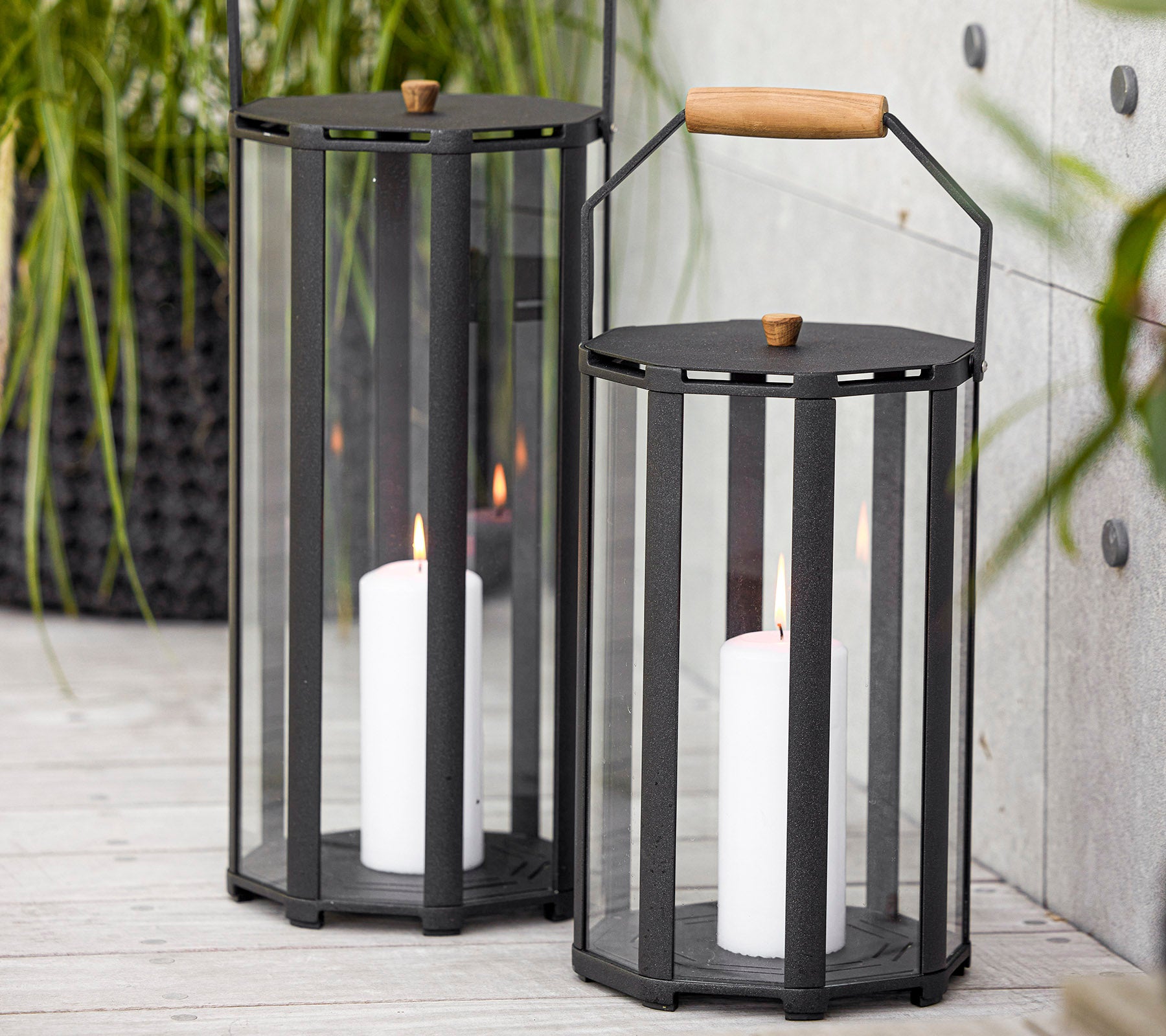 Dos faroles elegantes con un diseño moderno, que cuentan con paneles de vidrio y un mango de madera, sosteniendo velas blancas en su interior.