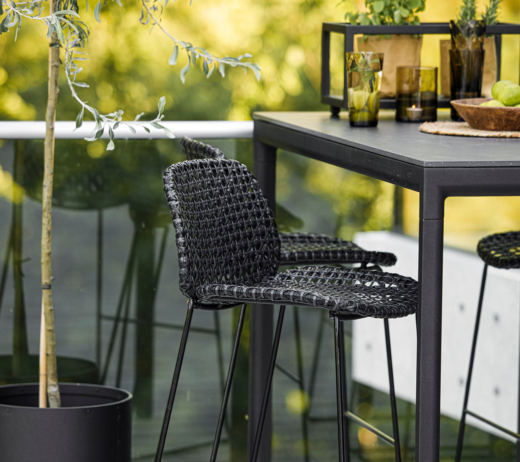 Una silla tejida negra elegante acompañada de una mesa elegante, rodeada de plantas decorativas y cristalería, creando un ambiente moderno.
