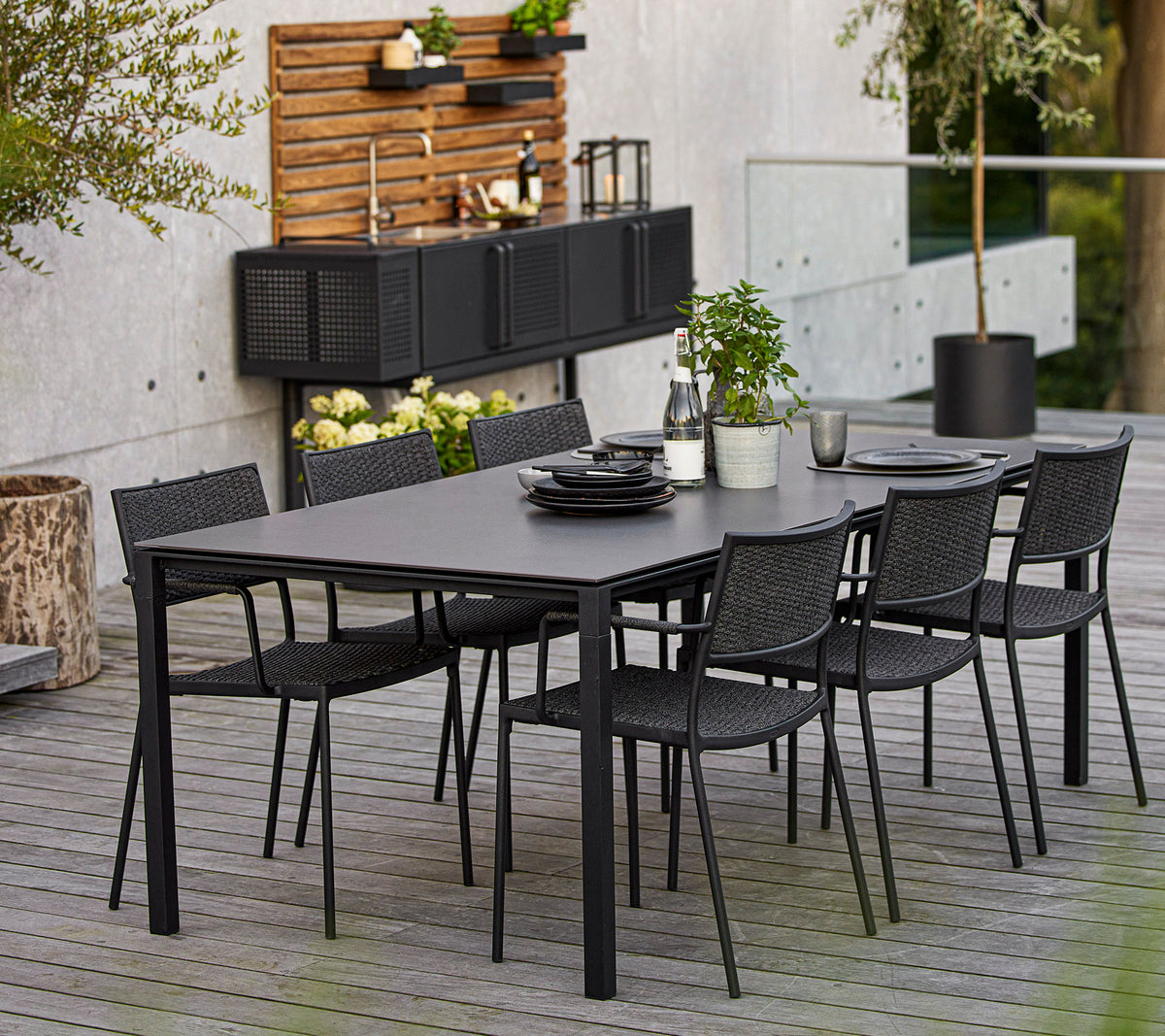 Un conjunto de comedor moderno que cuenta con una gran mesa rectangular y seis sillas de mimbre negras, acentuado con elementos decorativos y vegetación.