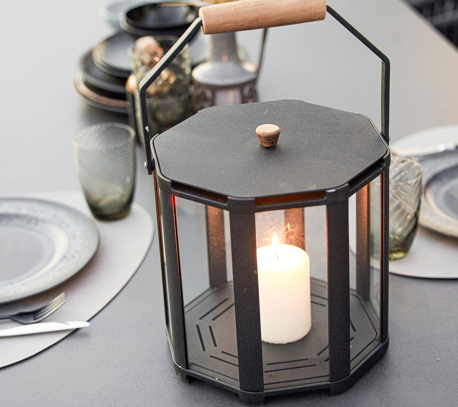Una elegante linterna octagonal con una vela central, rodeada de paneles de vidrio y una manija de madera, que realza cualquier ambiente.