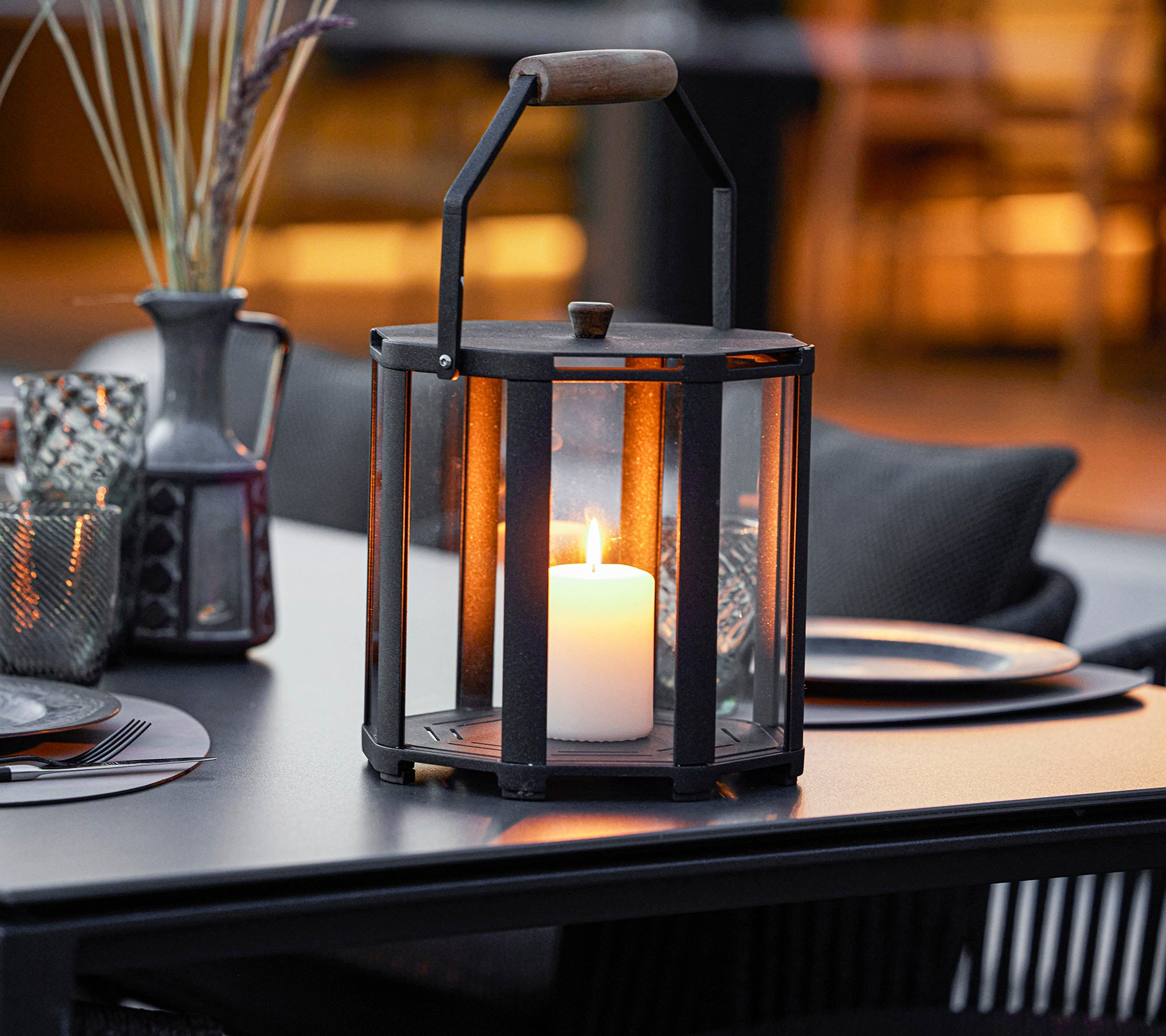 Una linterna elegante con una vela en su interior, con un marco de metal y un mango de madera, colocada sobre una mesa con elementos decorativos a su alrededor.