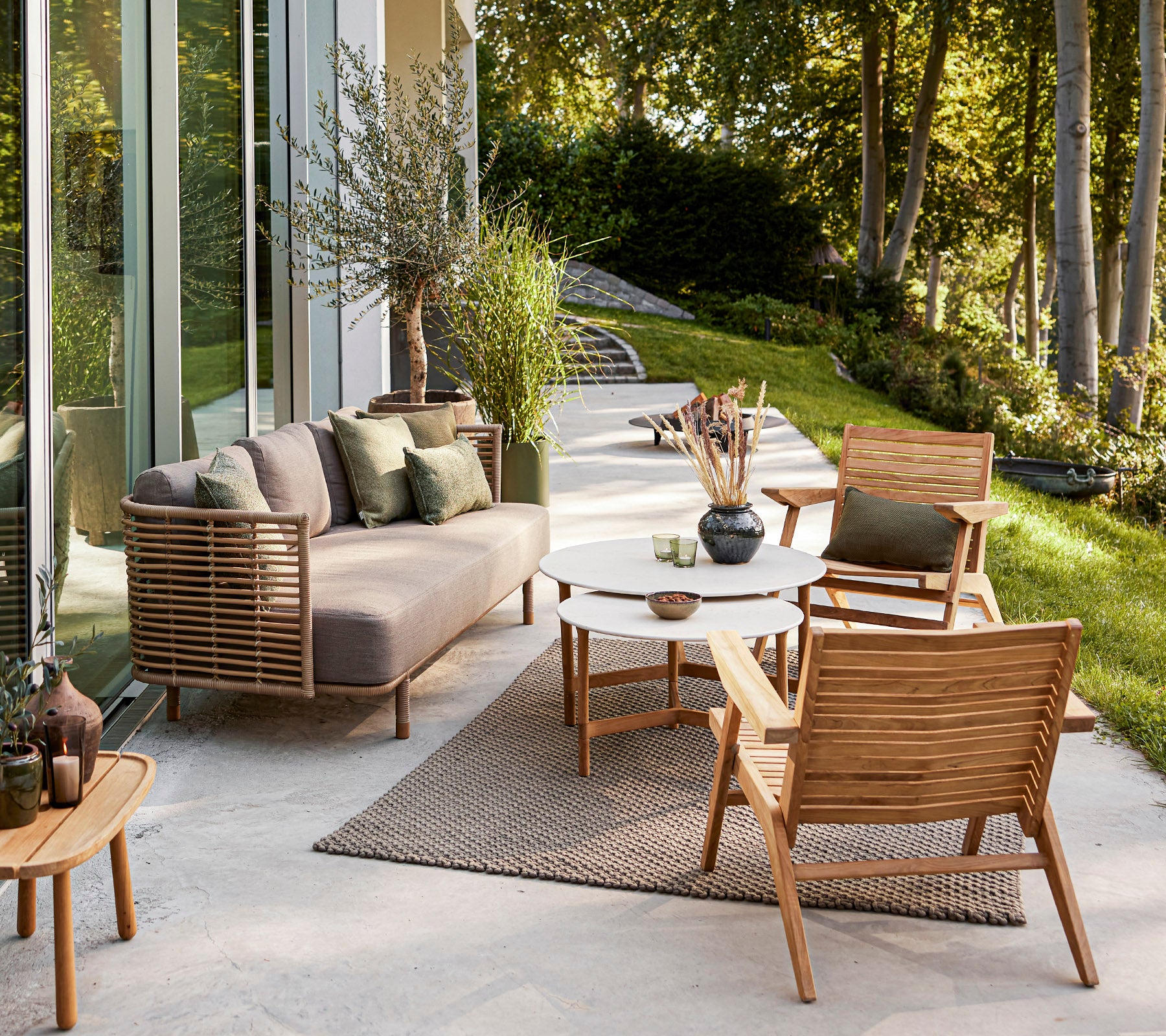 Área de estar al aire libre elegante con un sofá moderno, sillas de madera, una mesa redonda y plantas decorativas en un fondo sereno.