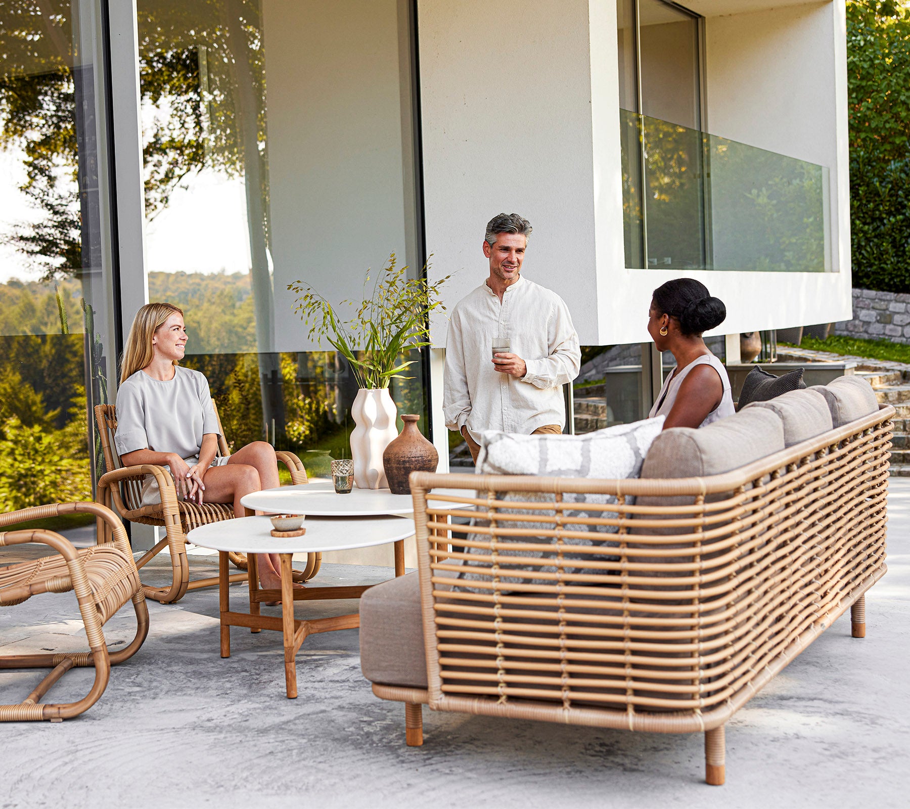 Tres personas disfrutan de una conversación informal sobre muebles de ratán elegantes con elementos decorativos, rodeados de vegetación.