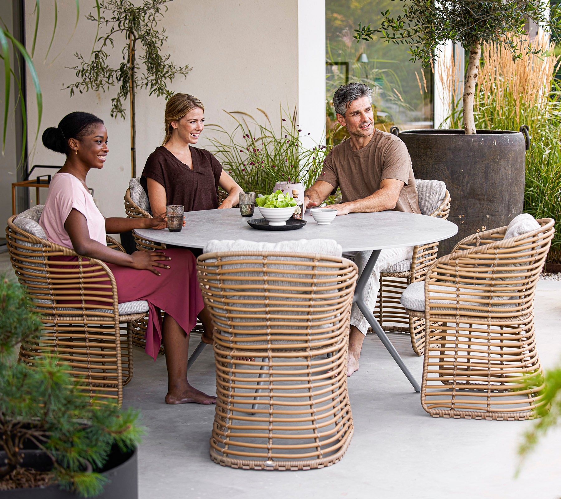 Cuatro personas disfrutan de bebidas alrededor de una mesa redonda moderna, con sillas de ratán elegantes y vegetación en un entorno sereno.