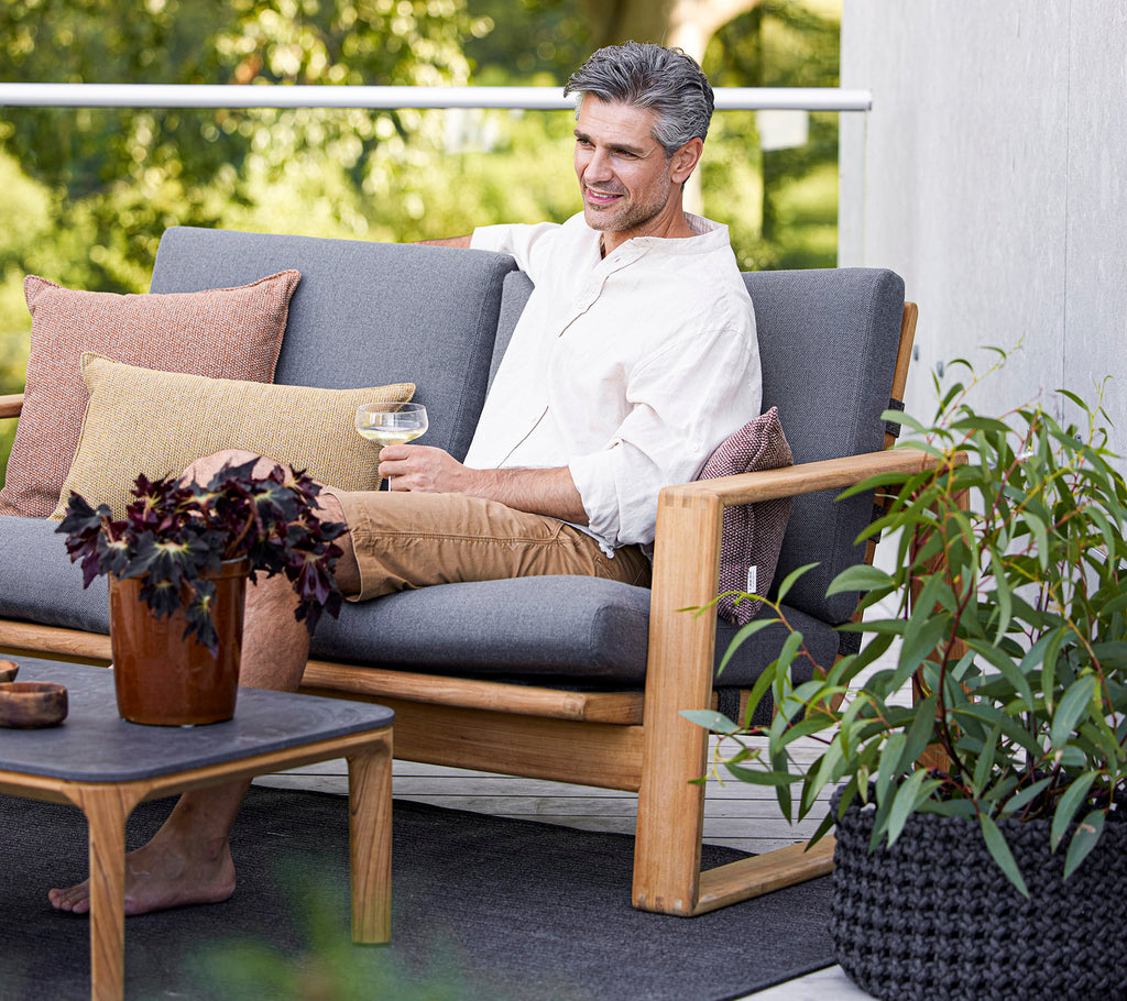 Un hombre relajado se sienta en un sofá elegante con cojines, disfrutando de una bebida junto a una planta en maceta y una mesa de café.