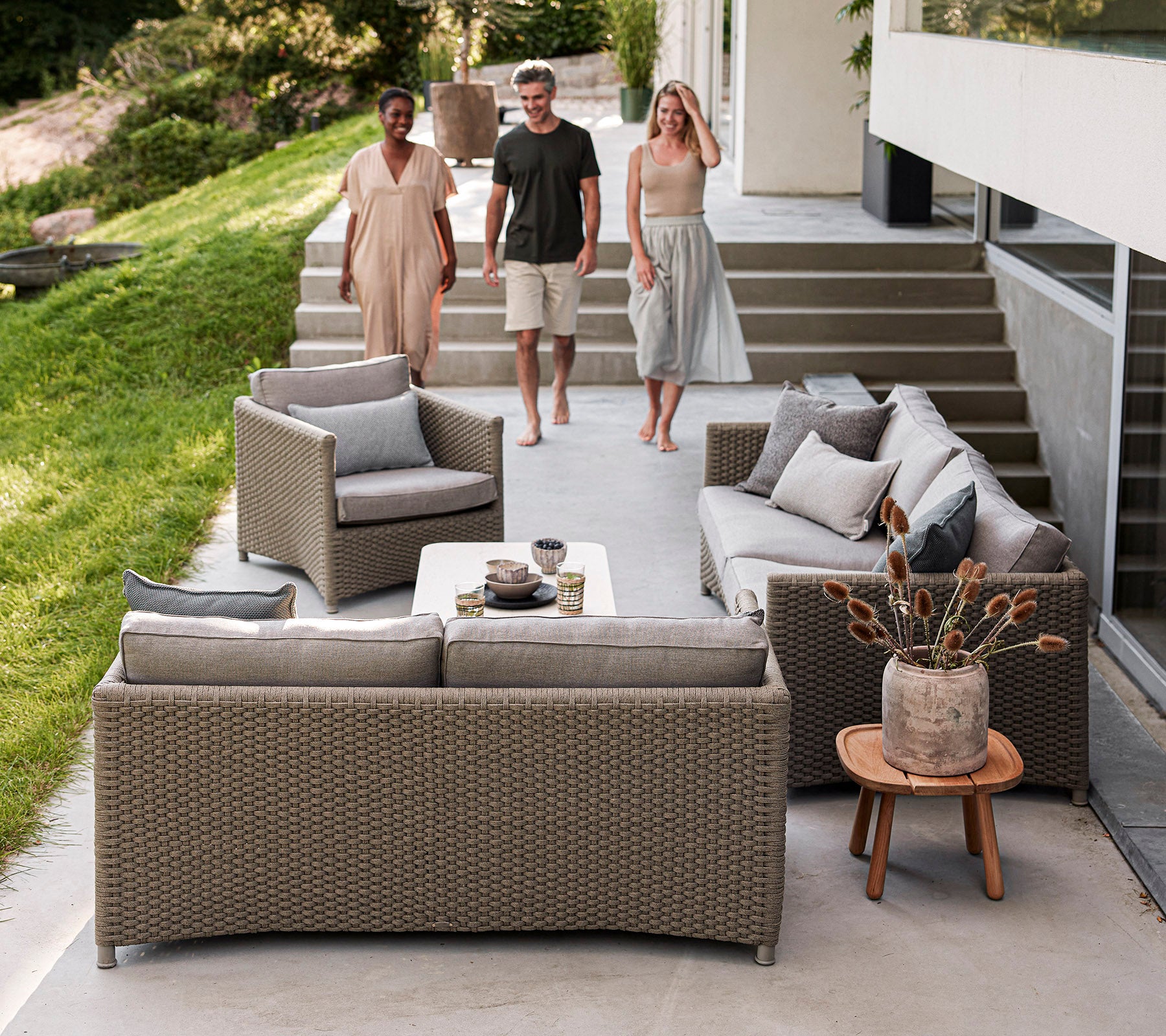 Tres personas caminan hacia un área de asientos al aire libre con estilo que cuenta con muebles de mimbre acogedores, cojines decorativos y una mesa de café.
