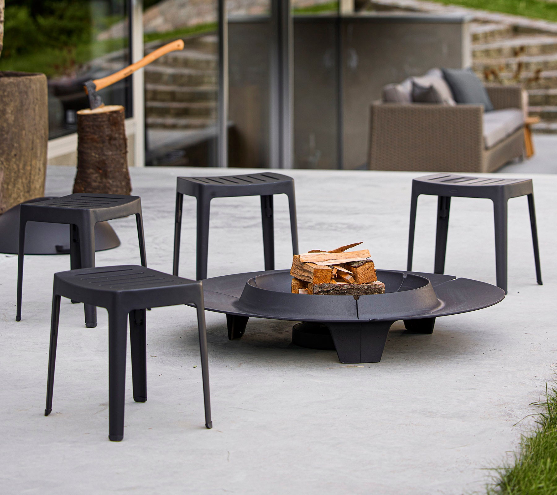 Una fogata circular rodeada de cuatro elegantes taburetes negros, con una pila de madera en el centro. Espacio moderno de reunión al aire libre.